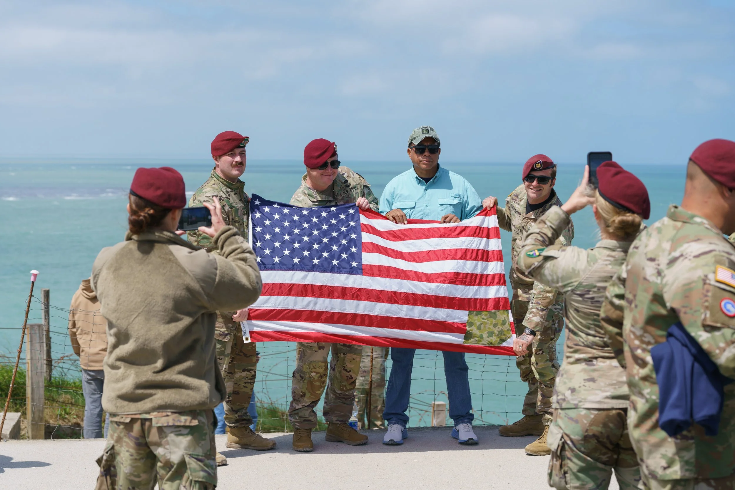 Military personnel and a civilian holding an American flag, photos being taken, near a body of water, with a cloudy sky.