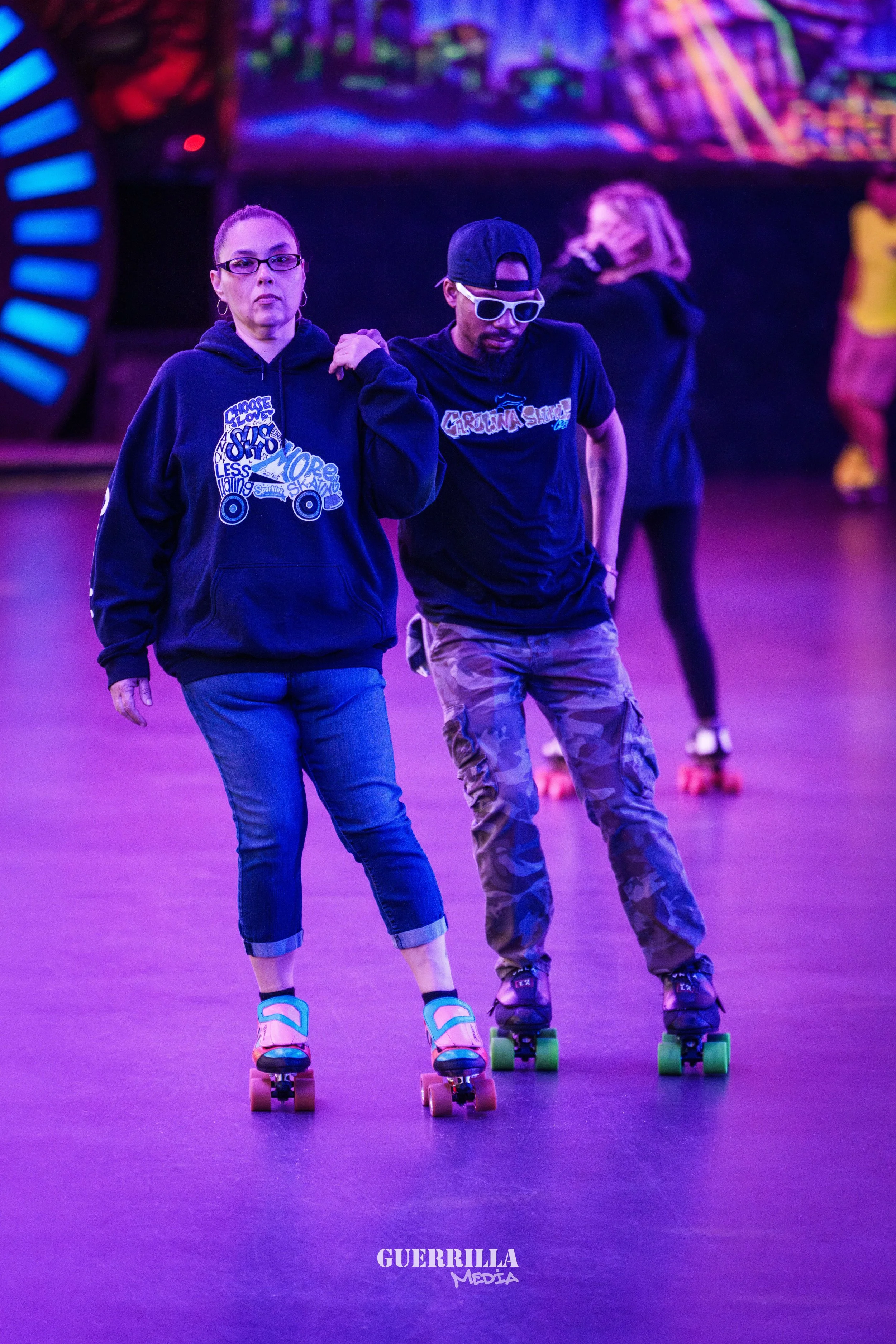 Two people roller skating in an indoor roller rink with purple lighting, one man and one woman holding onto each other's shoulders, with other skaters in the background.