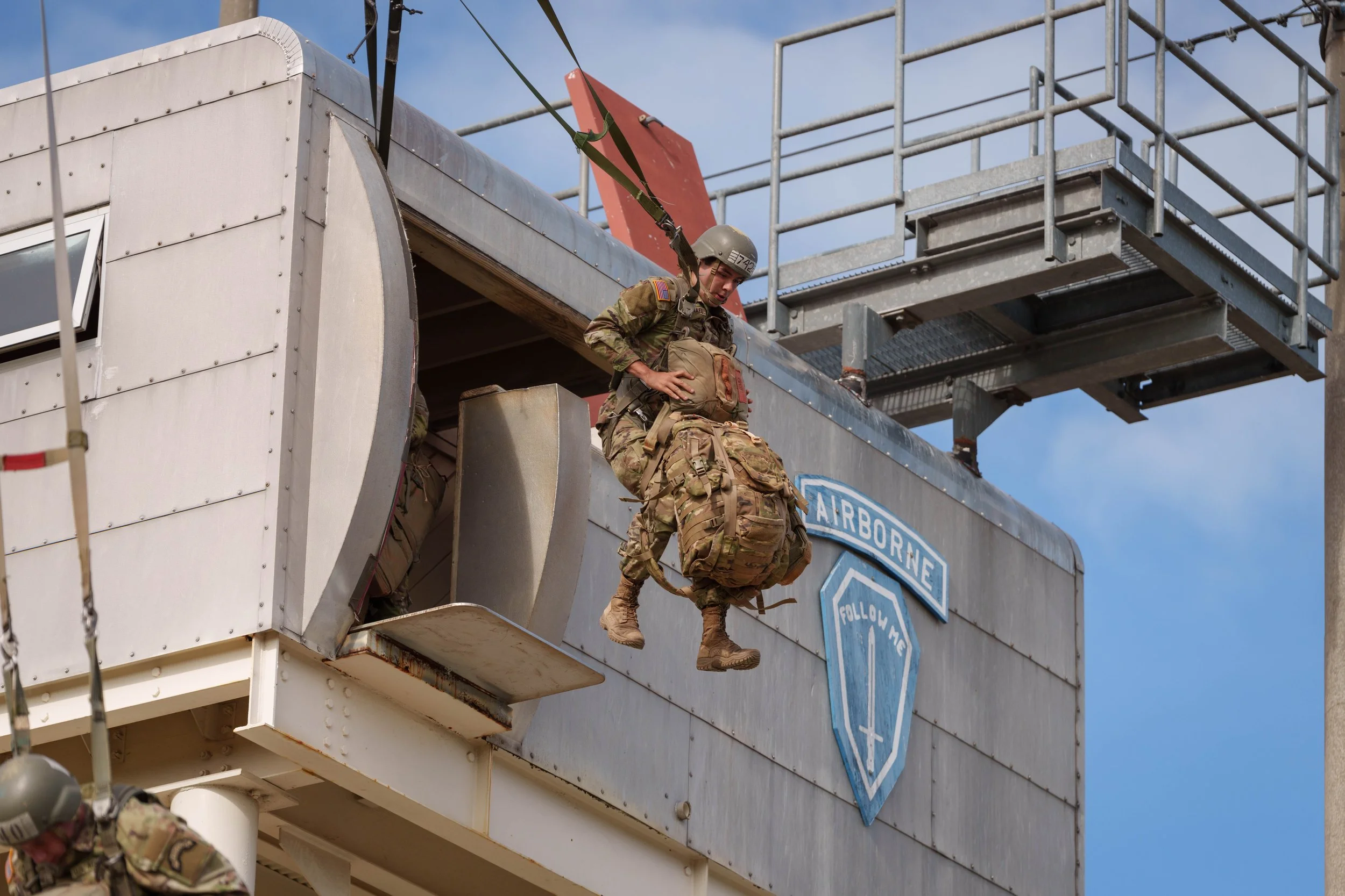 U.S. soldier in combat gear jumping out of an aircraft with an 'Airborne' patch, during a training exercise against a blue sky background.