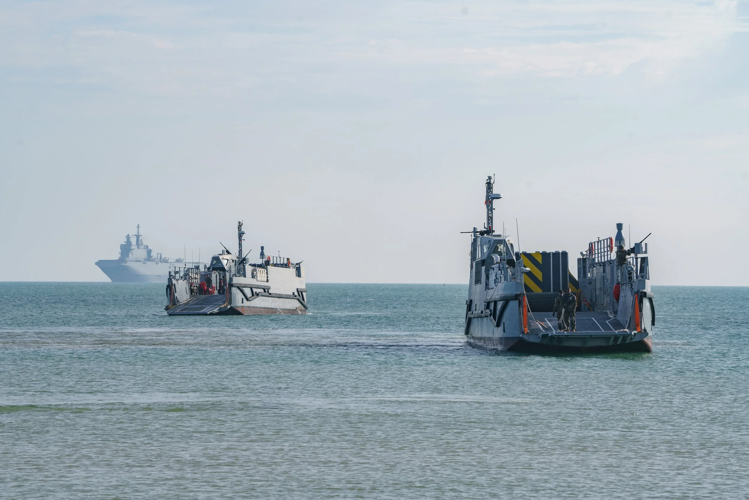 Two military boats approaching the shore with a larger ship visible in the distance on the ocean.