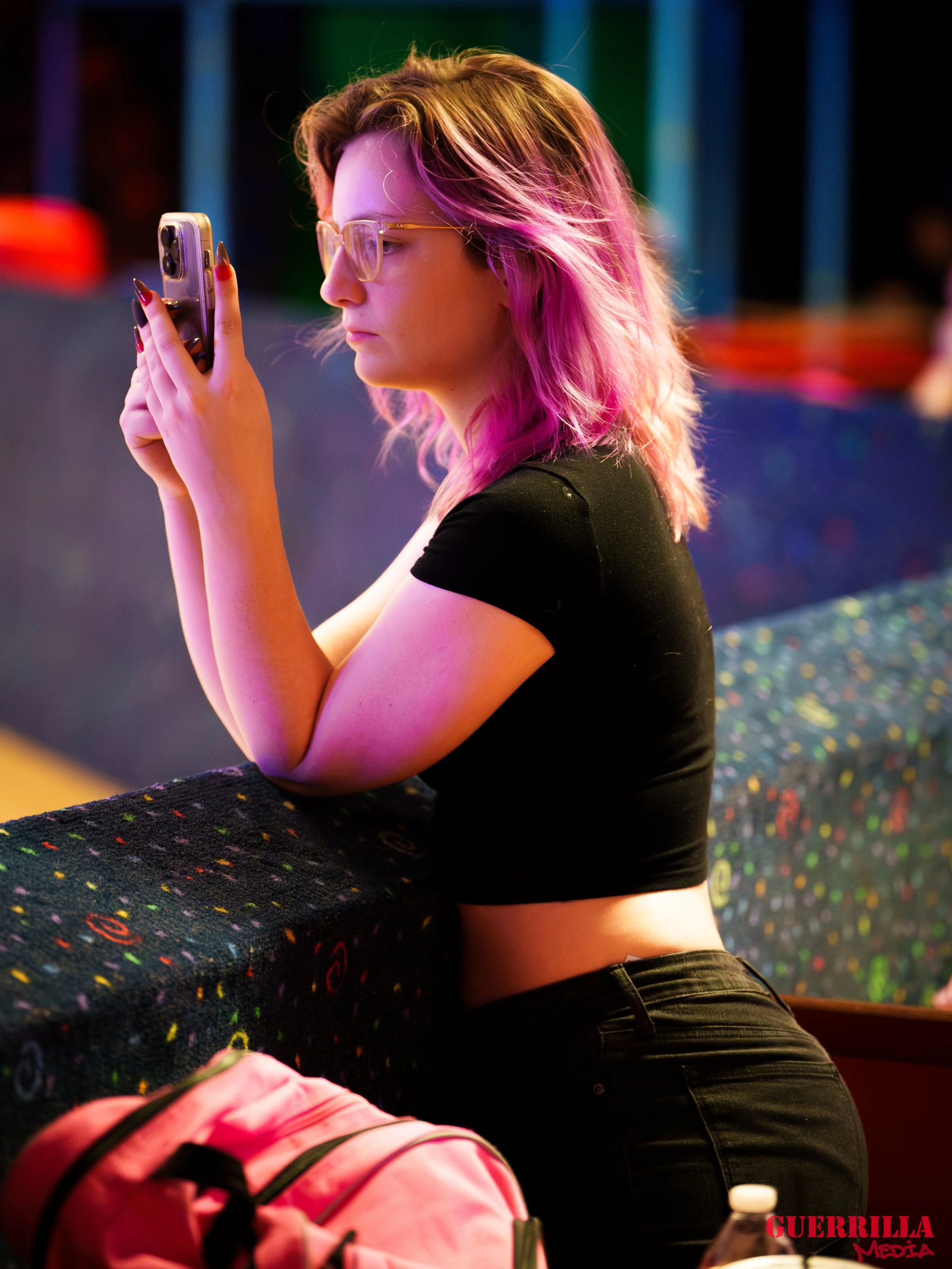 A young woman with pink hair, wearing glasses and a black crop top, stands at a railing in a colorful, lively setting while looking at her phone.