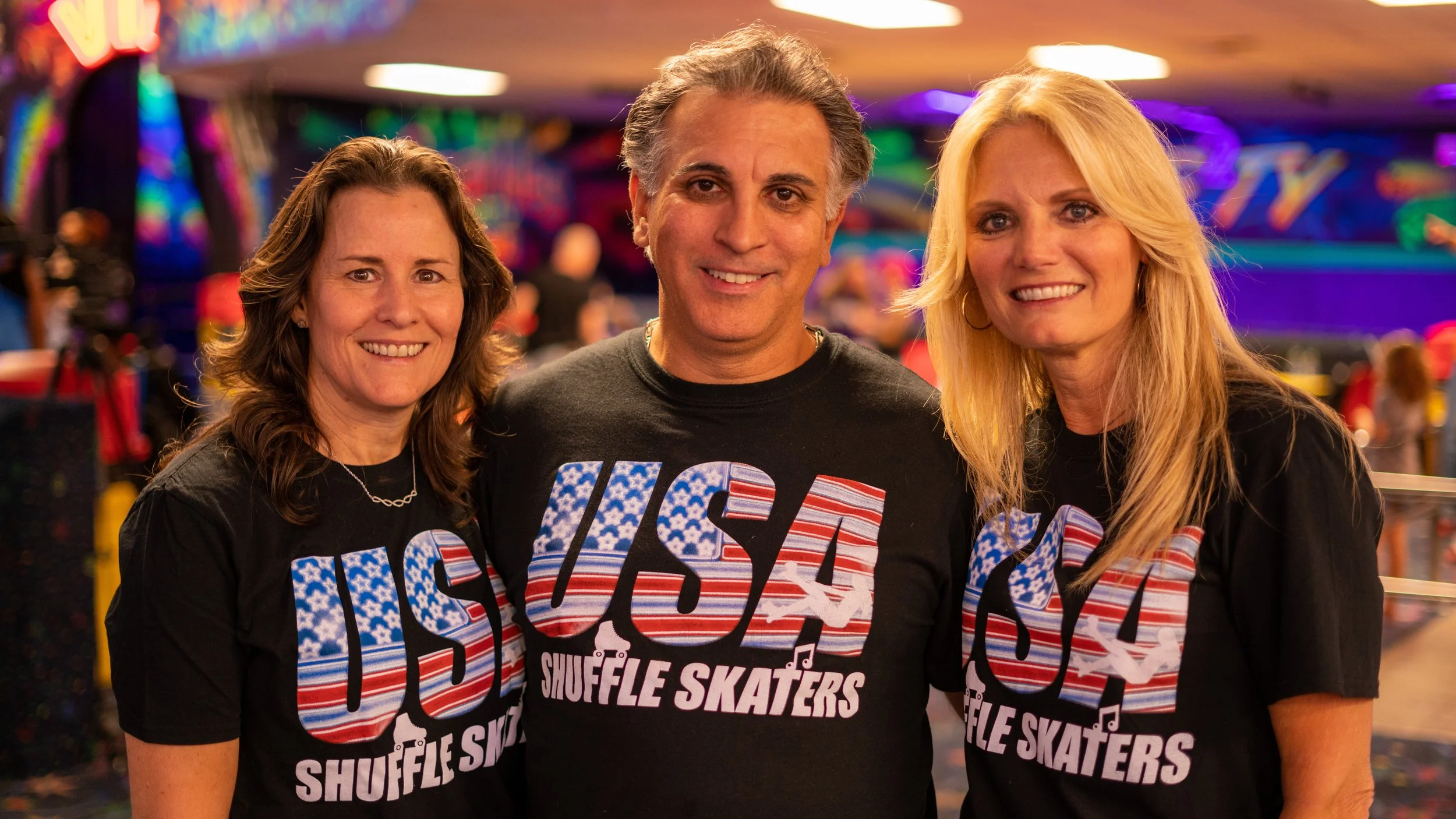 Three smiling people wearing black T-shirts with 'USA Shuffle Skaters' in patriotic colors, standing in an arcade or entertainment center with colorful lights and blurred background.