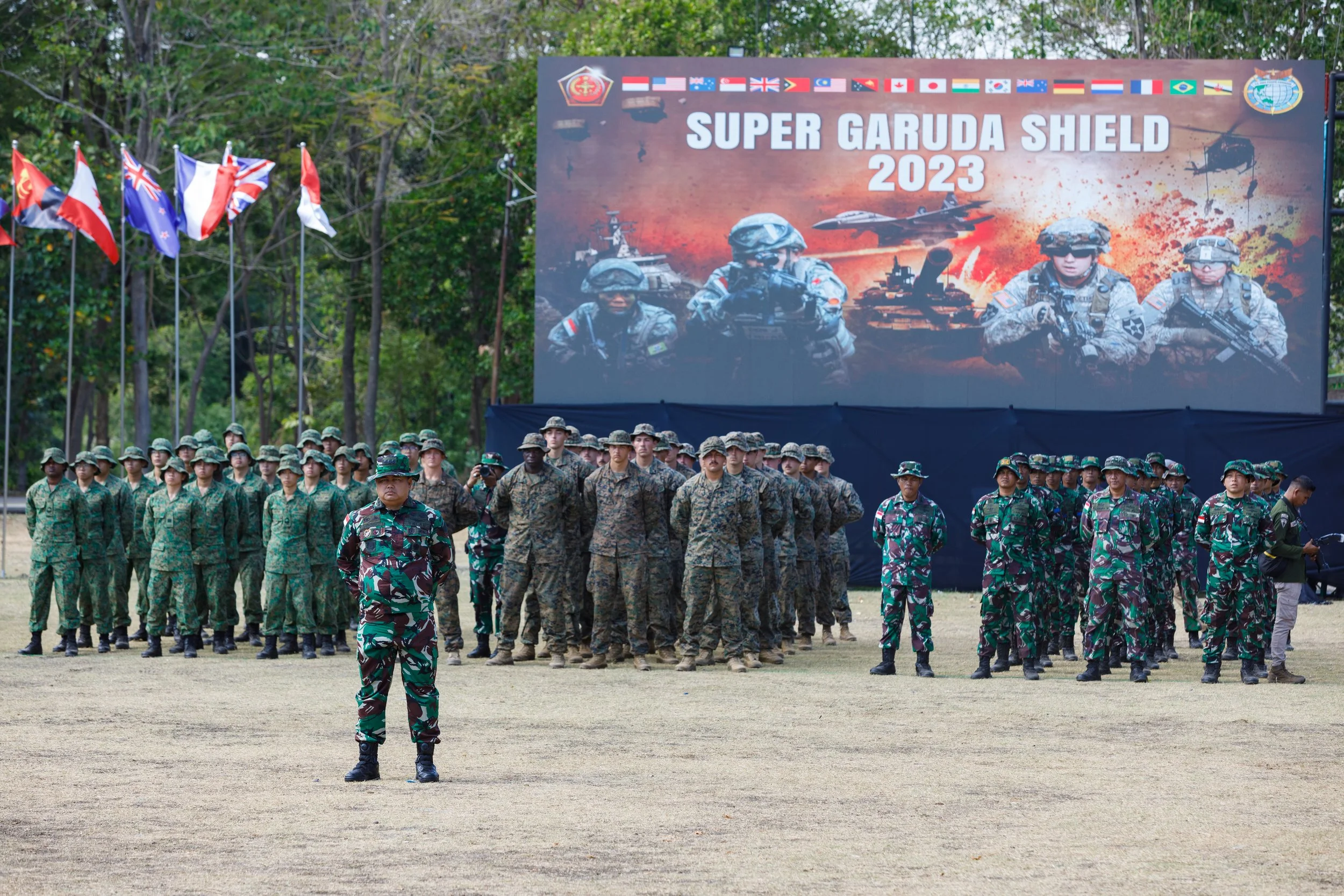 Military personnel stand in formation at an outdoor event, with flags and a large banner reading 'Super Garuda Shield 2023' in the background, featuring soldiers and military equipment.