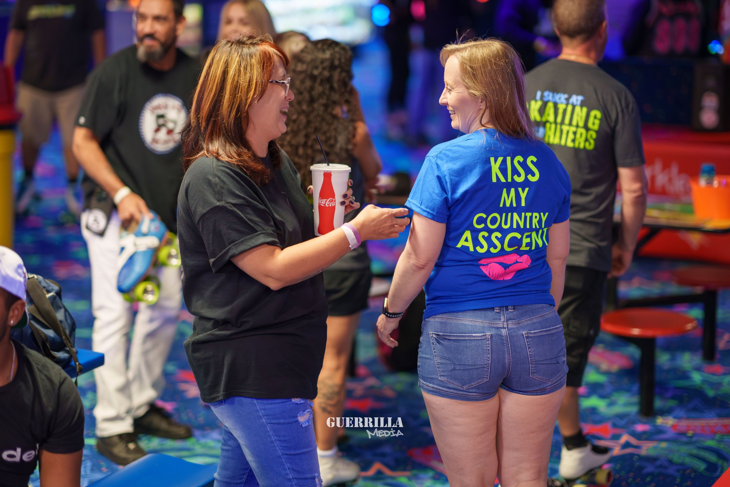Two women smiling and engaging in conversation at an indoor event. One woman with red hair is wearing a black shirt and holding a Coca-Cola cup, while the other woman with blonde hair is wearing a bright blue t-shirt with text and a lipstick graphic,