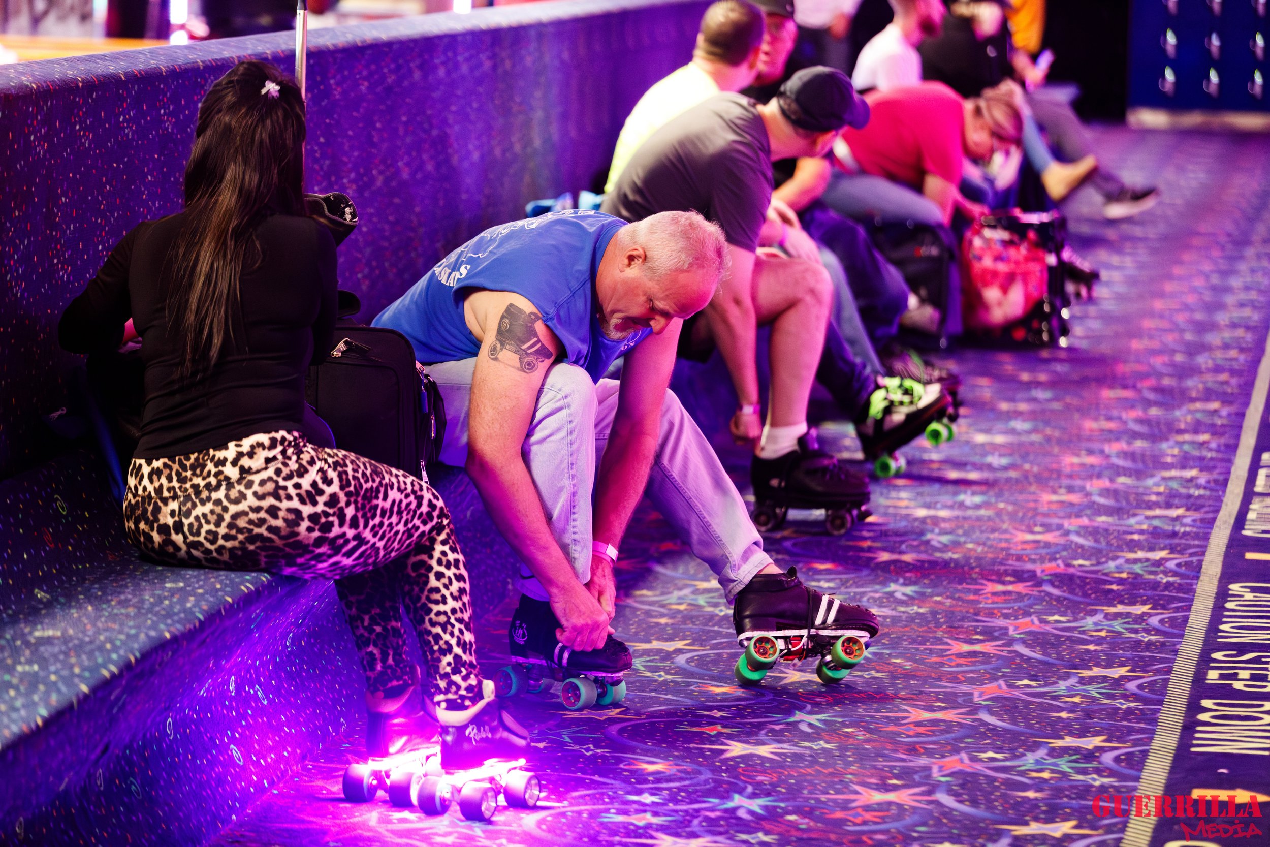 A group of people sitting on a bench at an indoor roller skating rink, putting on roller skates, with colorful neon lighting and star-patterned carpet.