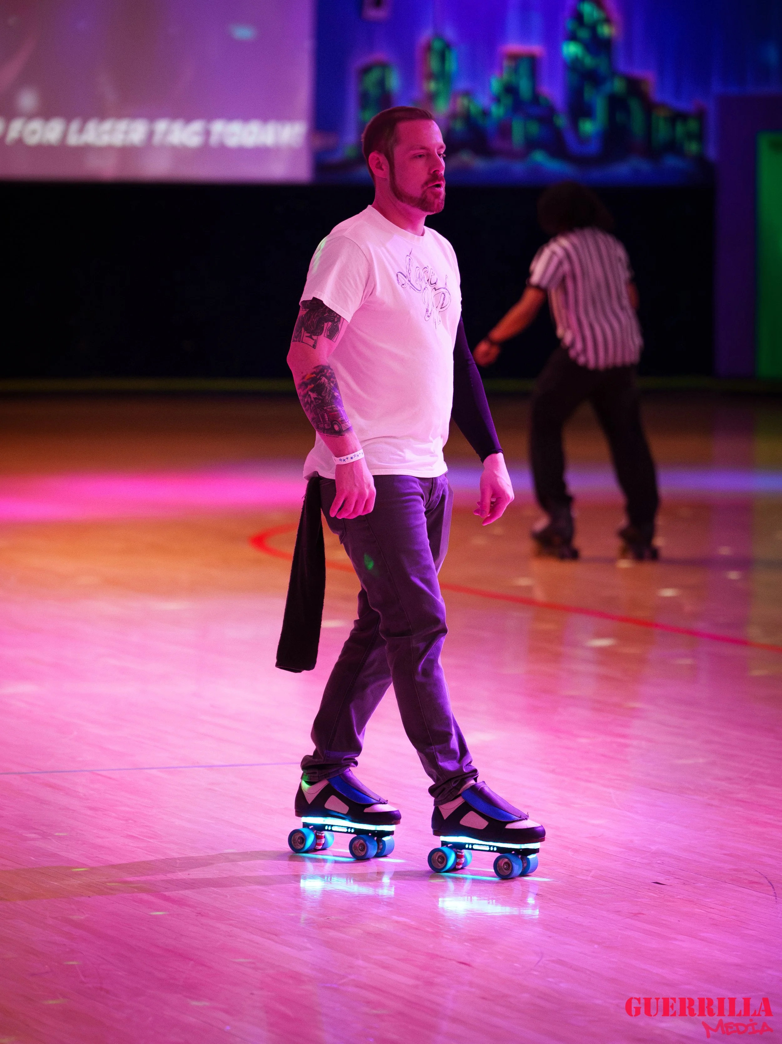 A man with tattoos on his arms wearing a white T-shirt and jeans roller skating on an indoor rink illuminated by pink and purple lights, with a woman in the background also roller skating.