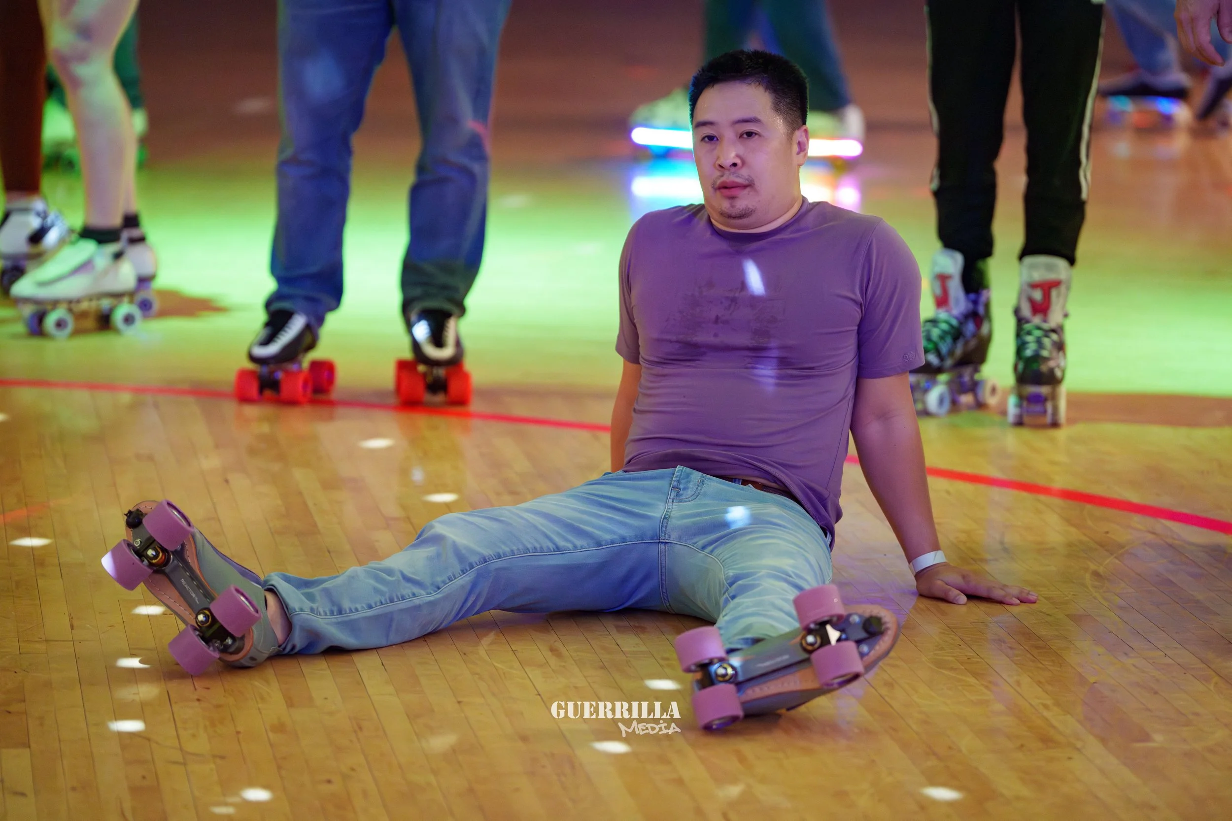 A man sitting on the floor of a roller skating rink, appearing tired or exhausted, with other skaters standing in the background.