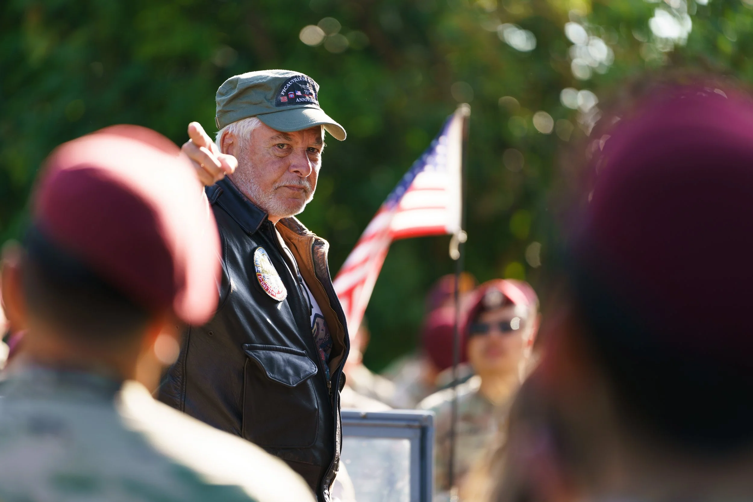 An older man with a gray beard wearing a cap and a leather vest speaking to a group of people outdoors, with an American flag in the background.