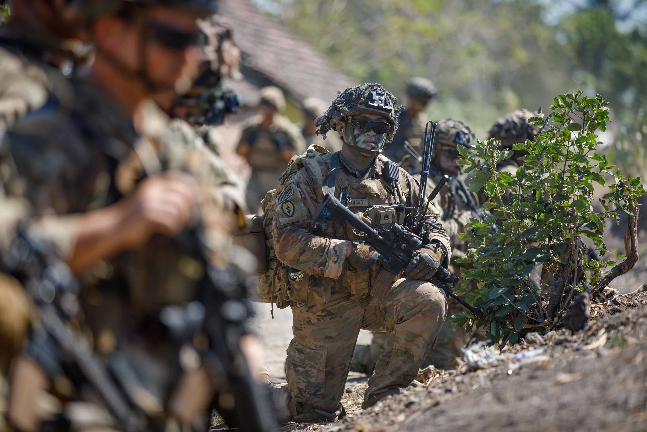Military personnel in camouflage uniforms and tactical gear kneeling and standing in a line outdoors during the daytime, with some holding rifles and others observing, amidst greenery and trees.