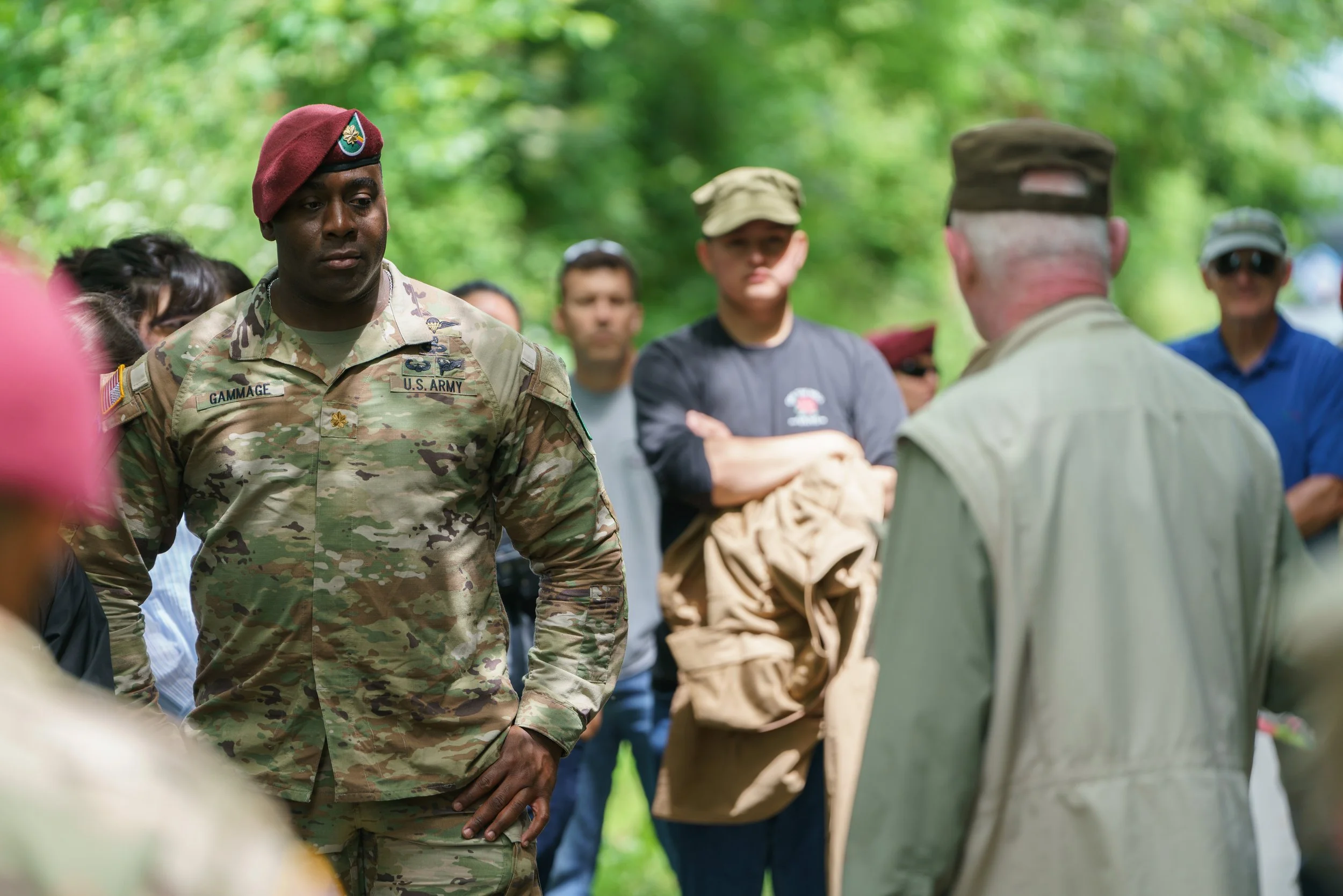 A U.S. Army soldier in camouflage uniform and a maroon beret standing with a group of people outdoors.