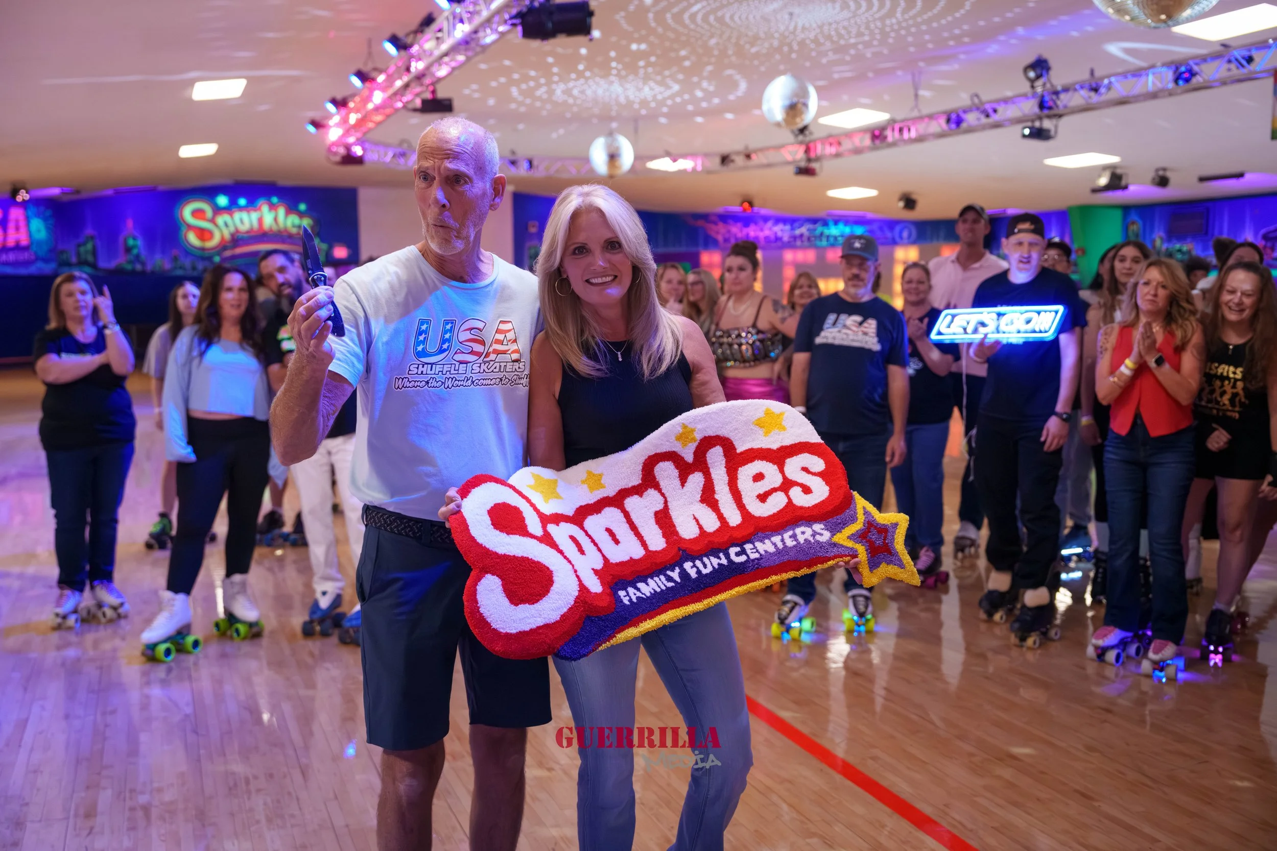 A group of people roller skating at a roller skating rink with colorful neon lights. Two people in the front hold a Sparklers sign, one man with a knife and a woman smiling. Many people are in the background, some holding signs and others skating.