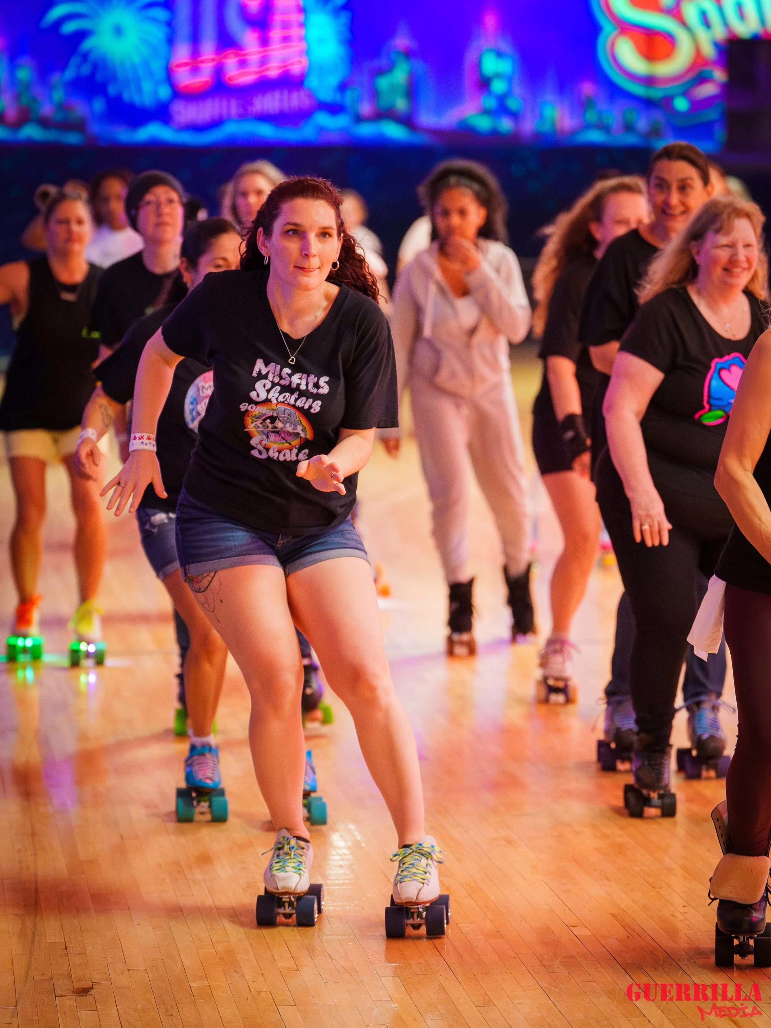 A group of people roller skating indoors on a wooden floor with a colorful, neon-lit backdrop.