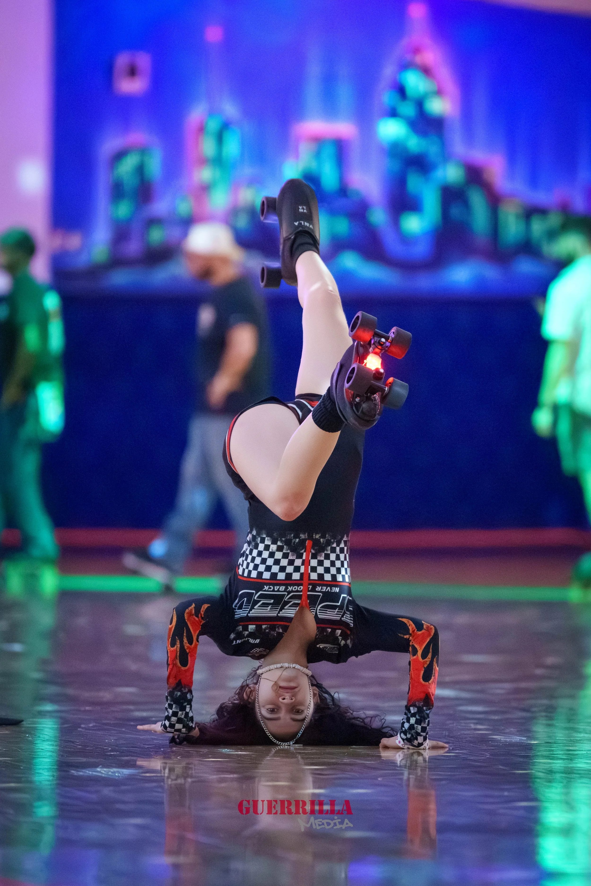 A young girl performing a headstand on the floor at a roller skating rink, wearing roller skates, a checkered and flame-themed outfit, with colorful neon lights in the background.
