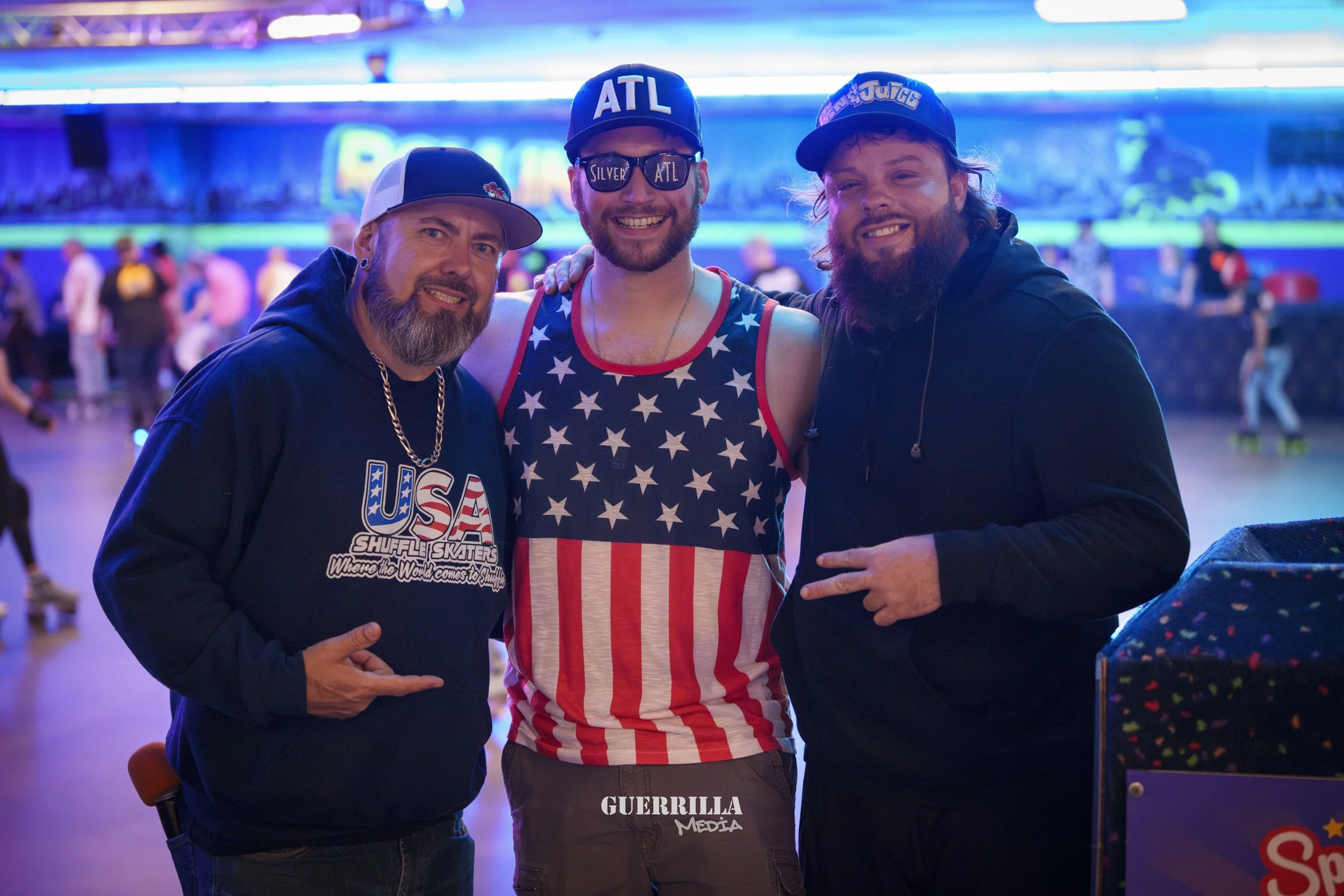 Three men standing together and smiling in an indoor roller skating rink with colorful lights, wearing casual clothing and caps.