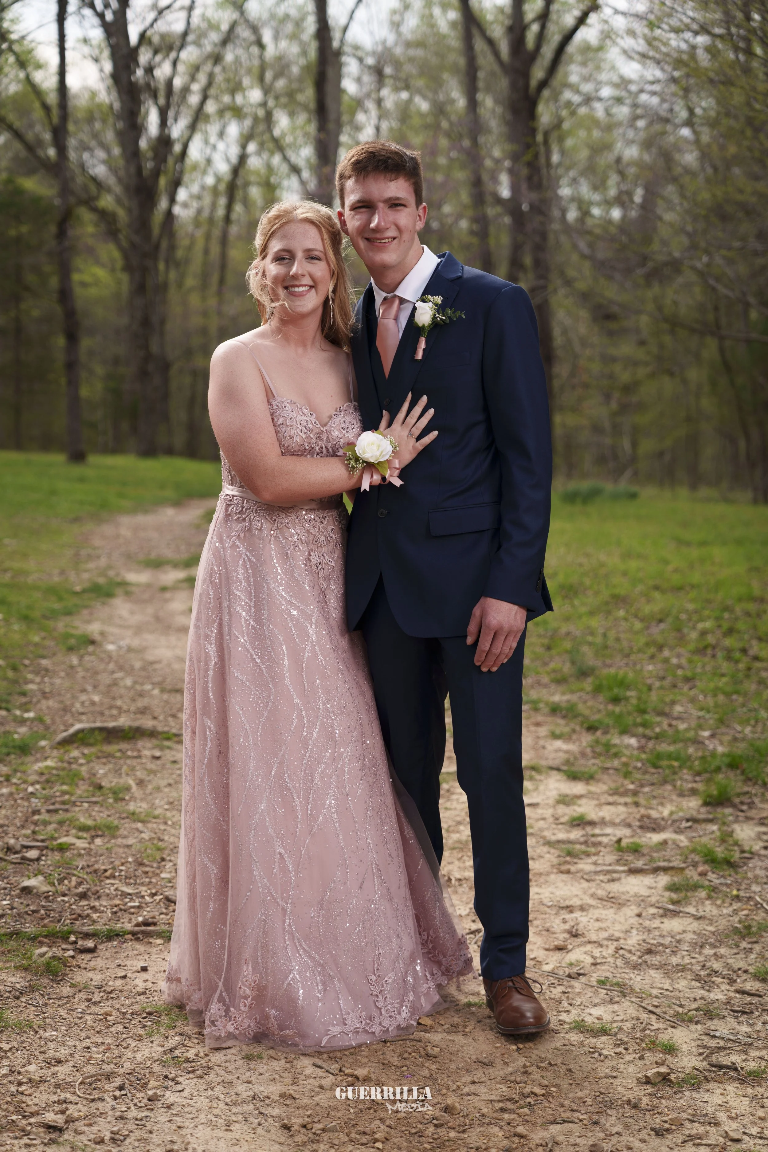 A young couple dressed in formal attire standing outdoors on a dirt path in a wooded area. The woman wears a pink, strapless gown with glittery embroidery, and the man wears a navy suit with a white shirt and light pink tie. They are smiling and hold