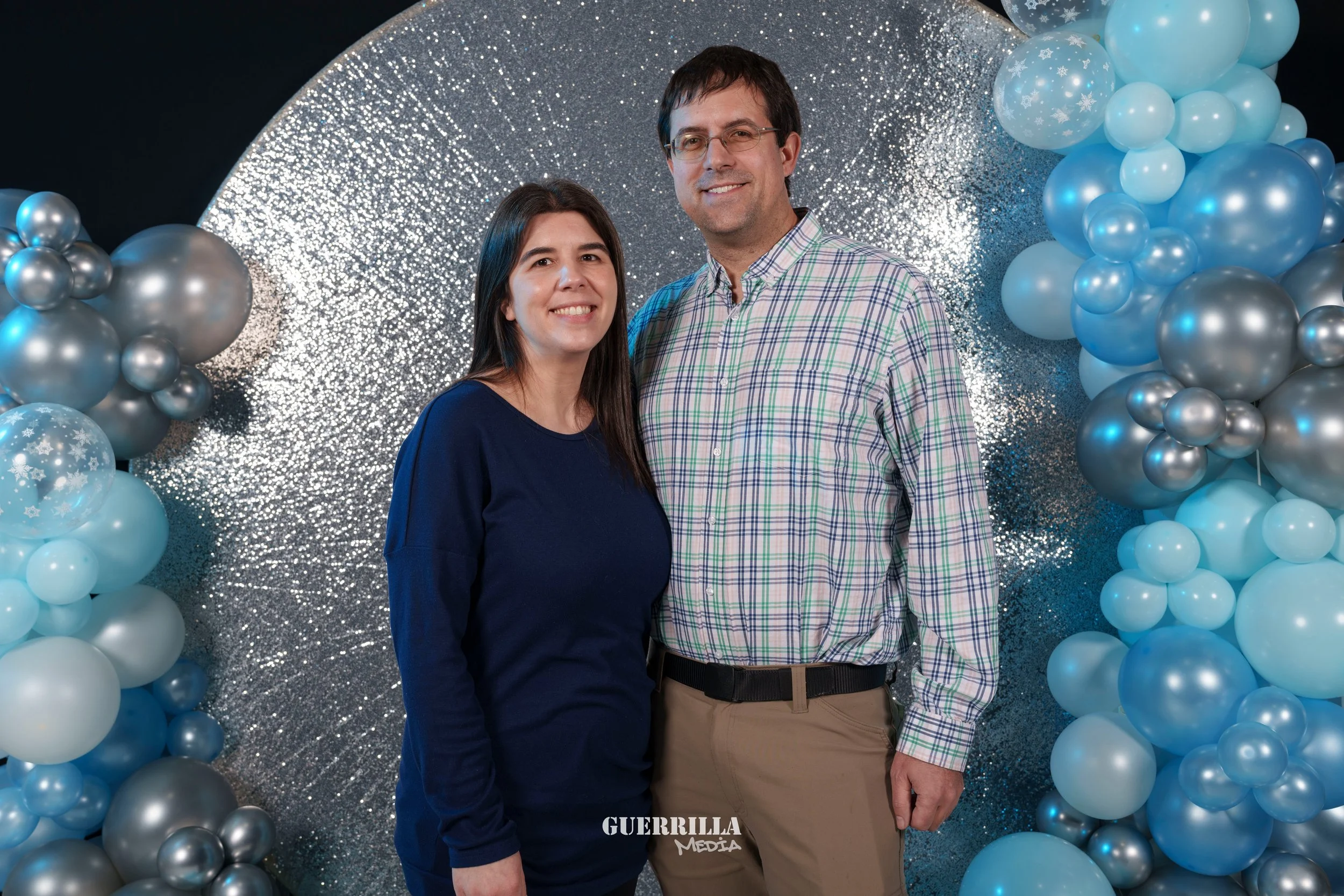A woman and a man standing in front of a silver glittery backdrop decorated with blue and silver balloons, smiling at the camera.