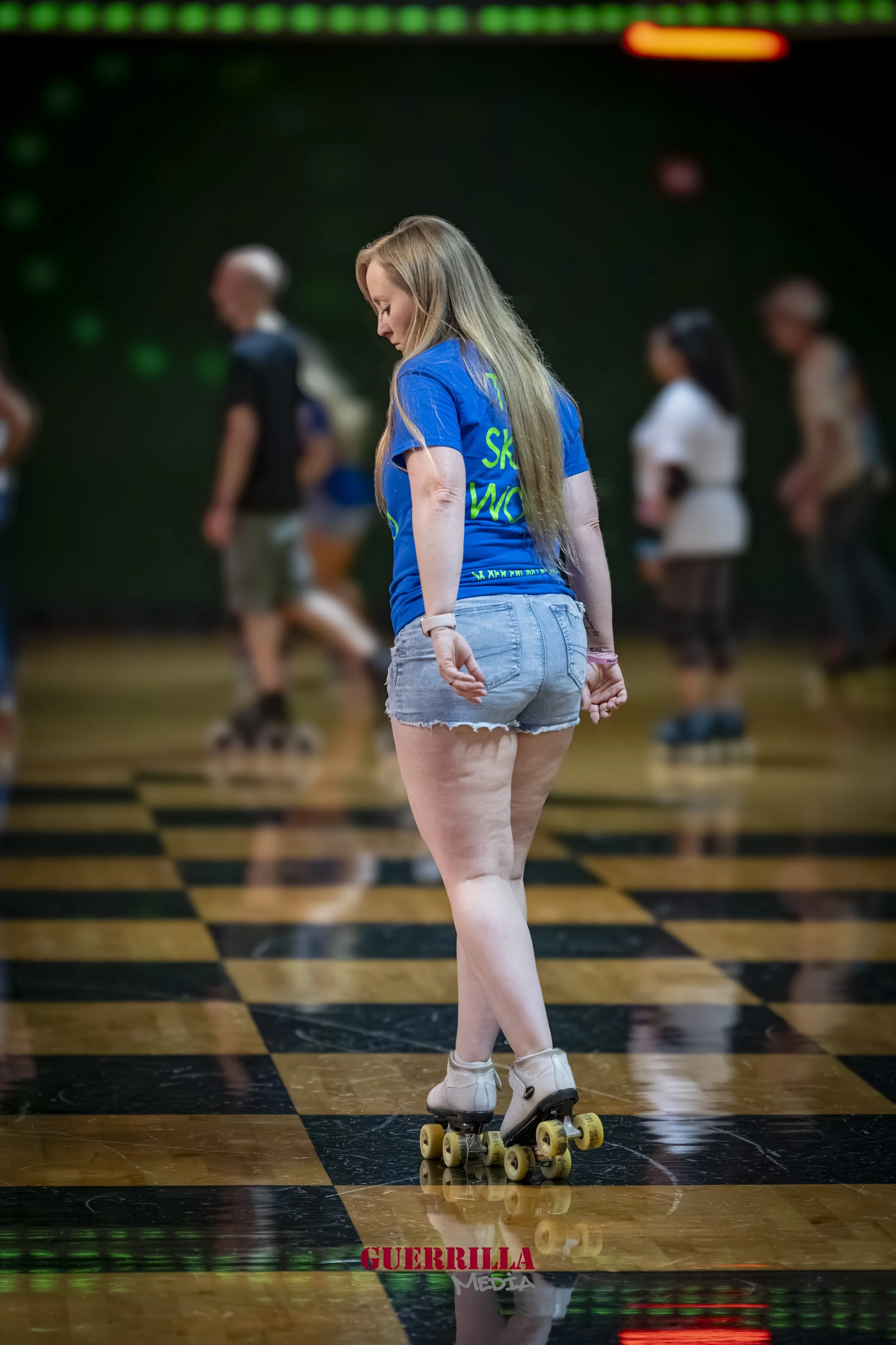 A woman roller skating on a checkered floor with multiple people in the background, some blurred, in an indoor skating rink.