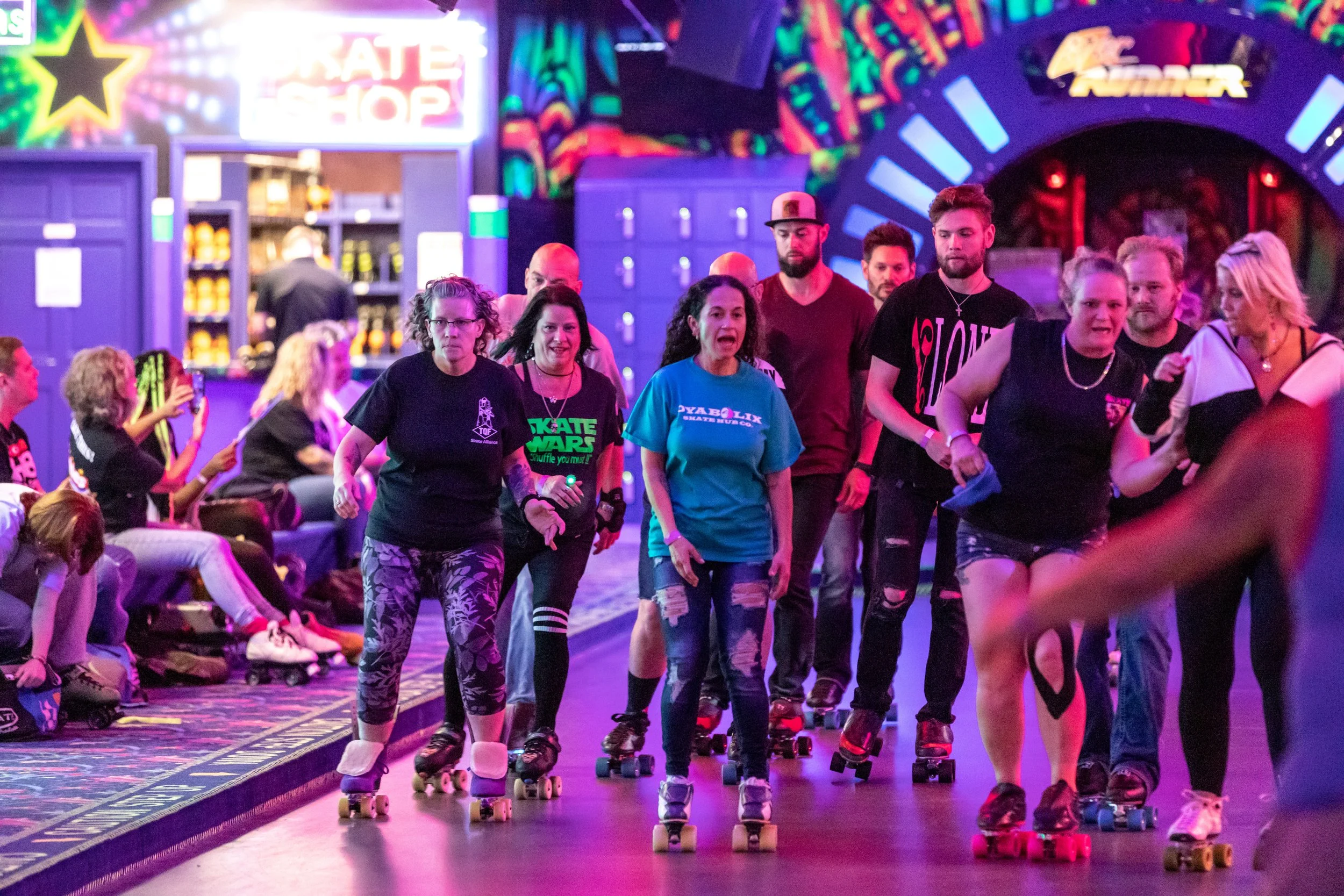 A group of people roller skating inside an arcade or roller rink, with colorful neon lights and advertisements in the background.