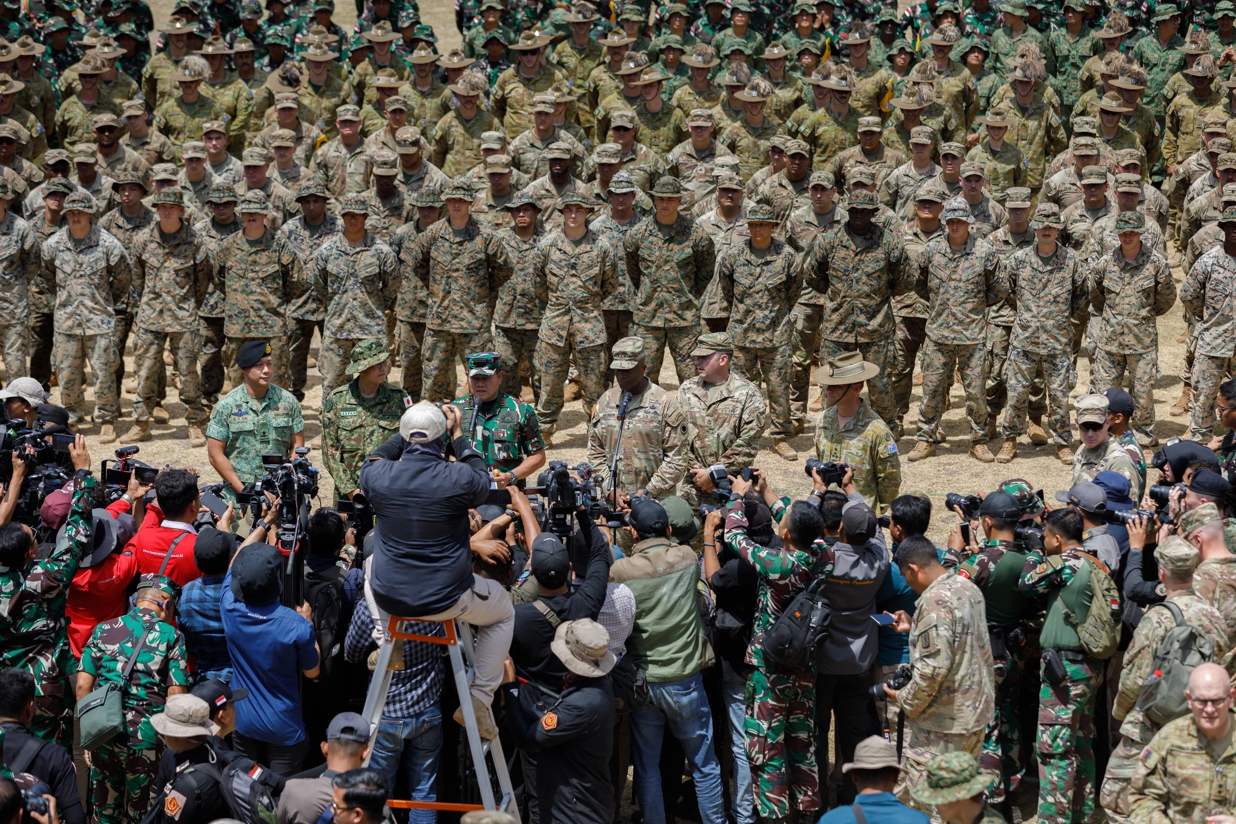 Military personnel gathering for a training or ceremony, with many soldiers standing in formation in camouflage uniforms and hats, while a group of journalists and photographers in civilian clothes and military uniforms take pictures and record the e