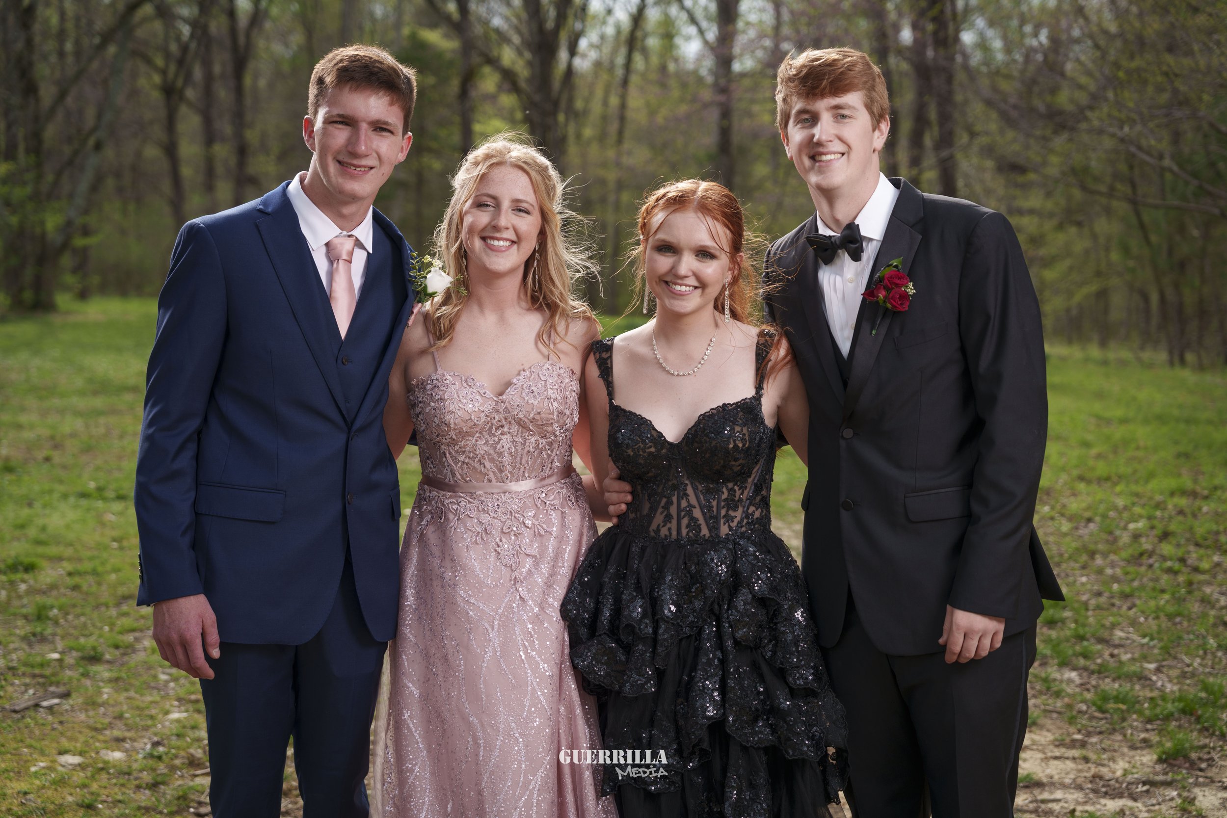 Four teenagers in formal attire are standing outdoors in a wooded area, smiling at the camera. Two boys are wearing tuxedos with boutonnieres, and two girls are wearing evening gowns, one in pink and one in black.