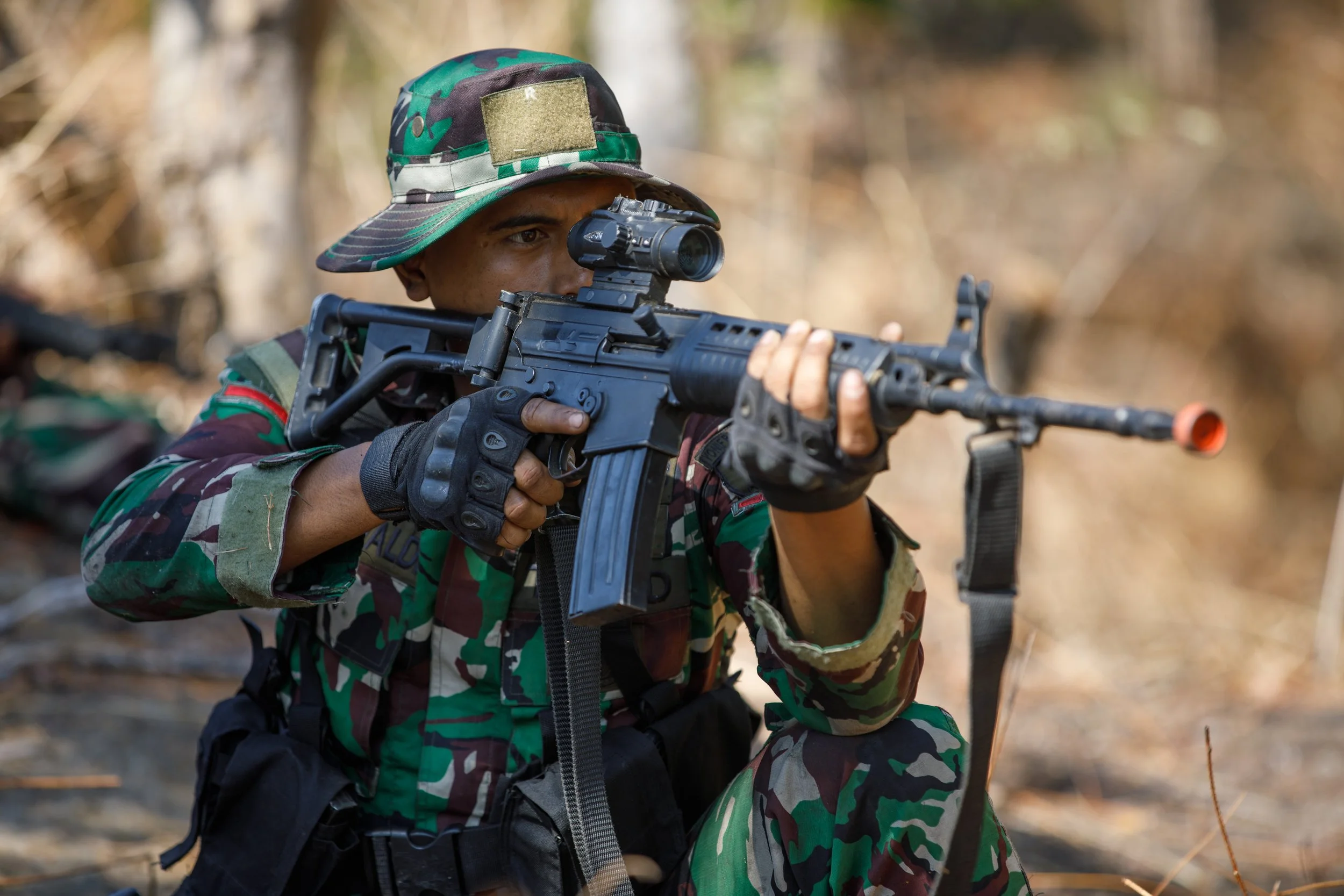 A soldier in camouflage uniform aiming a rifle with a scope in a wooded area.