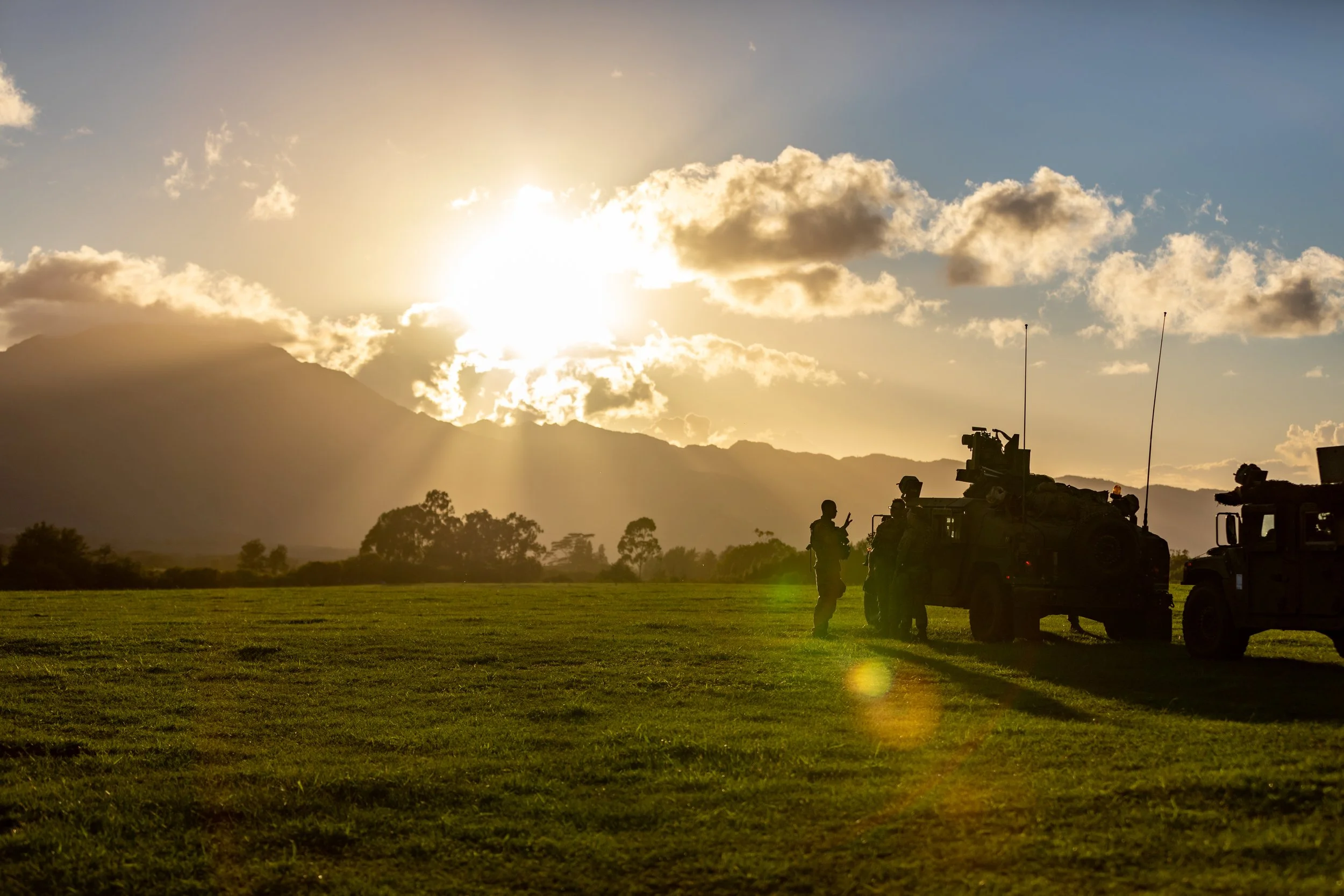 Soldiers standing near military vehicles on a grassy field at sunset with mountain backdrop.