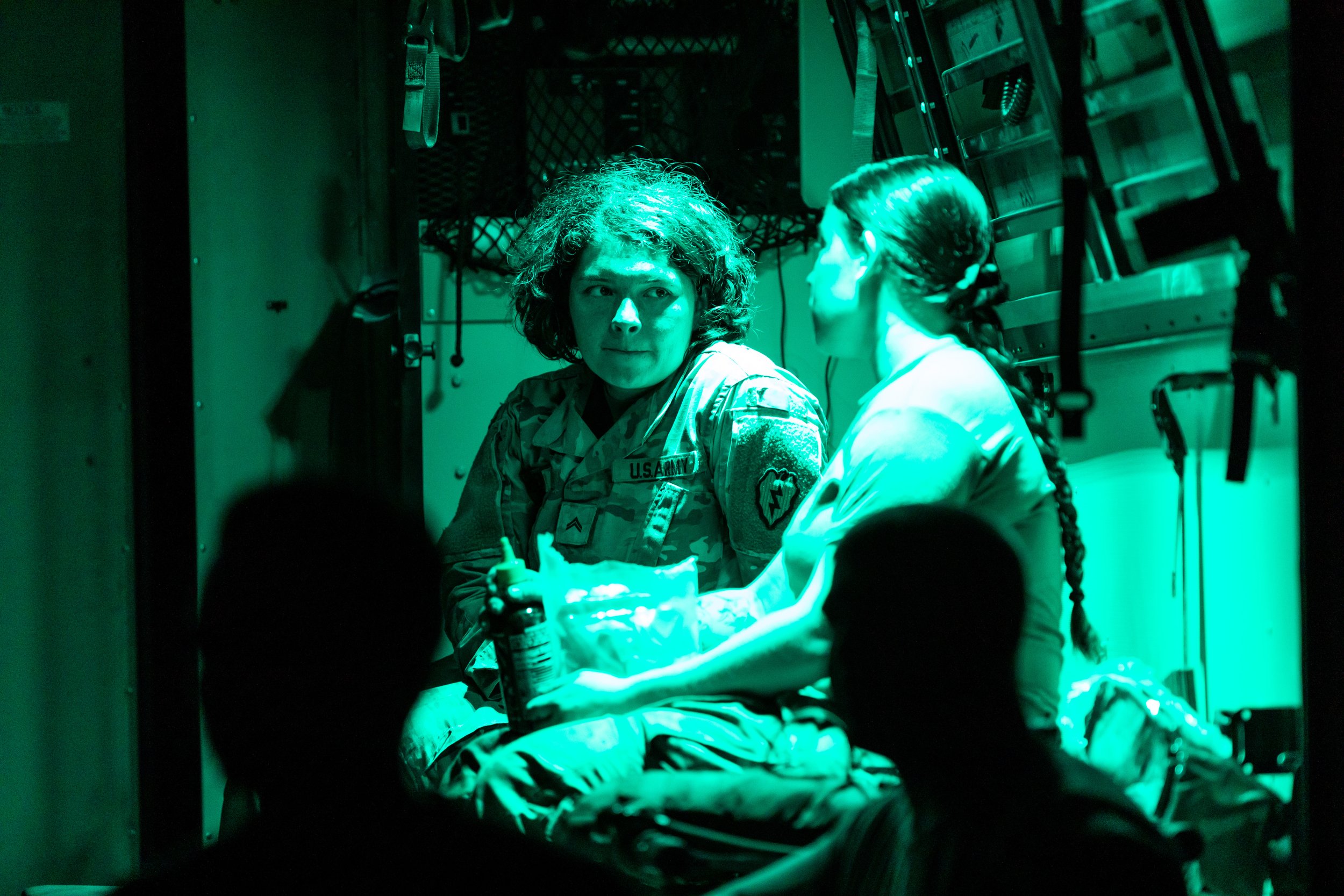 Two women sitting in a dimly lit room, one in military uniform and the other in a medical outfit, engaged in conversation. The room has military gear hanging on the walls and medical supplies around.