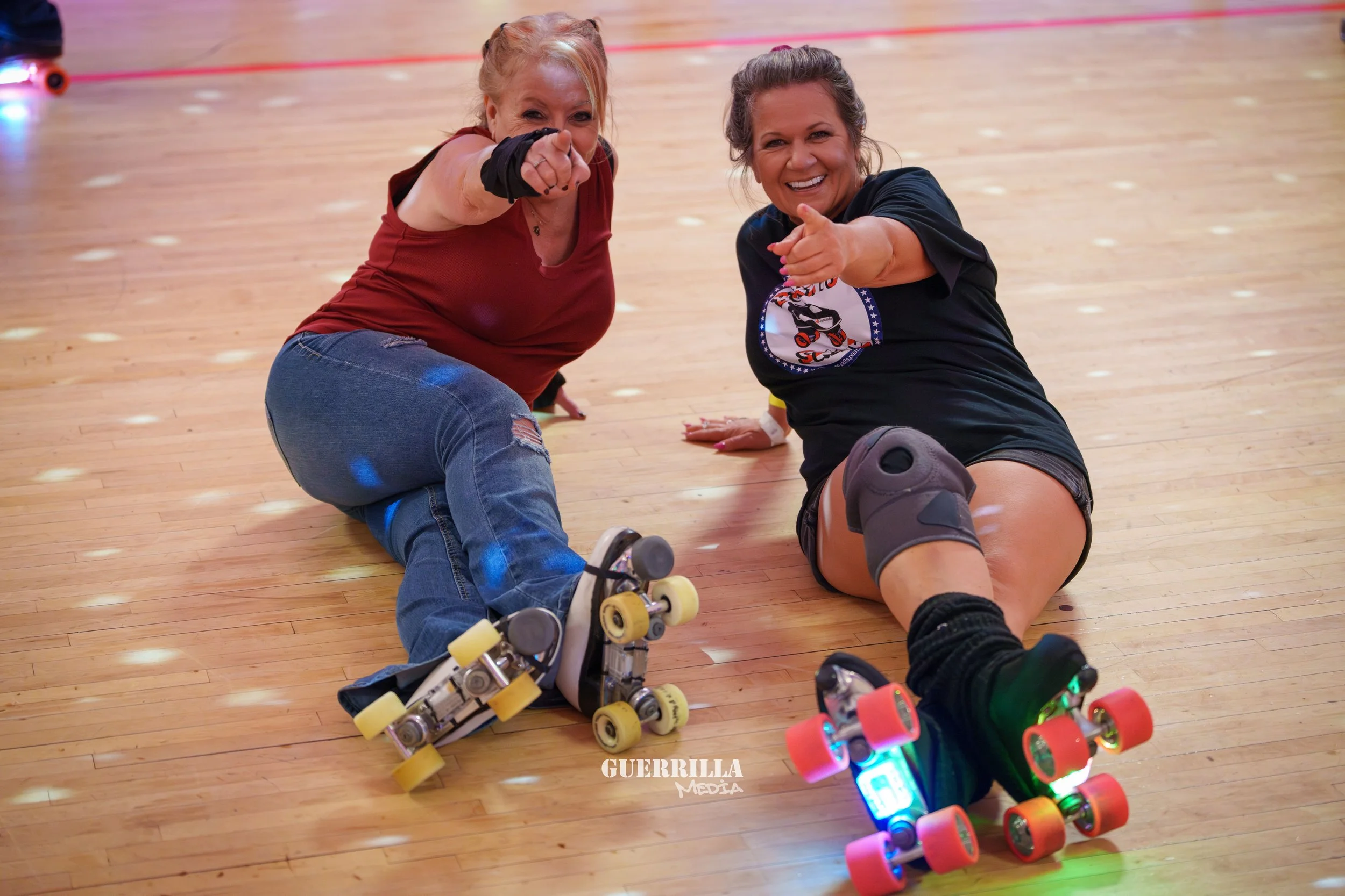 Two women on a roller skating rink sitting on the floor, pointing and smiling. They have roller skates on, and one is wearing a black t-shirt with a graphic and a knee pad, while the other is in a red tank top and ripped jeans. The background shows a
