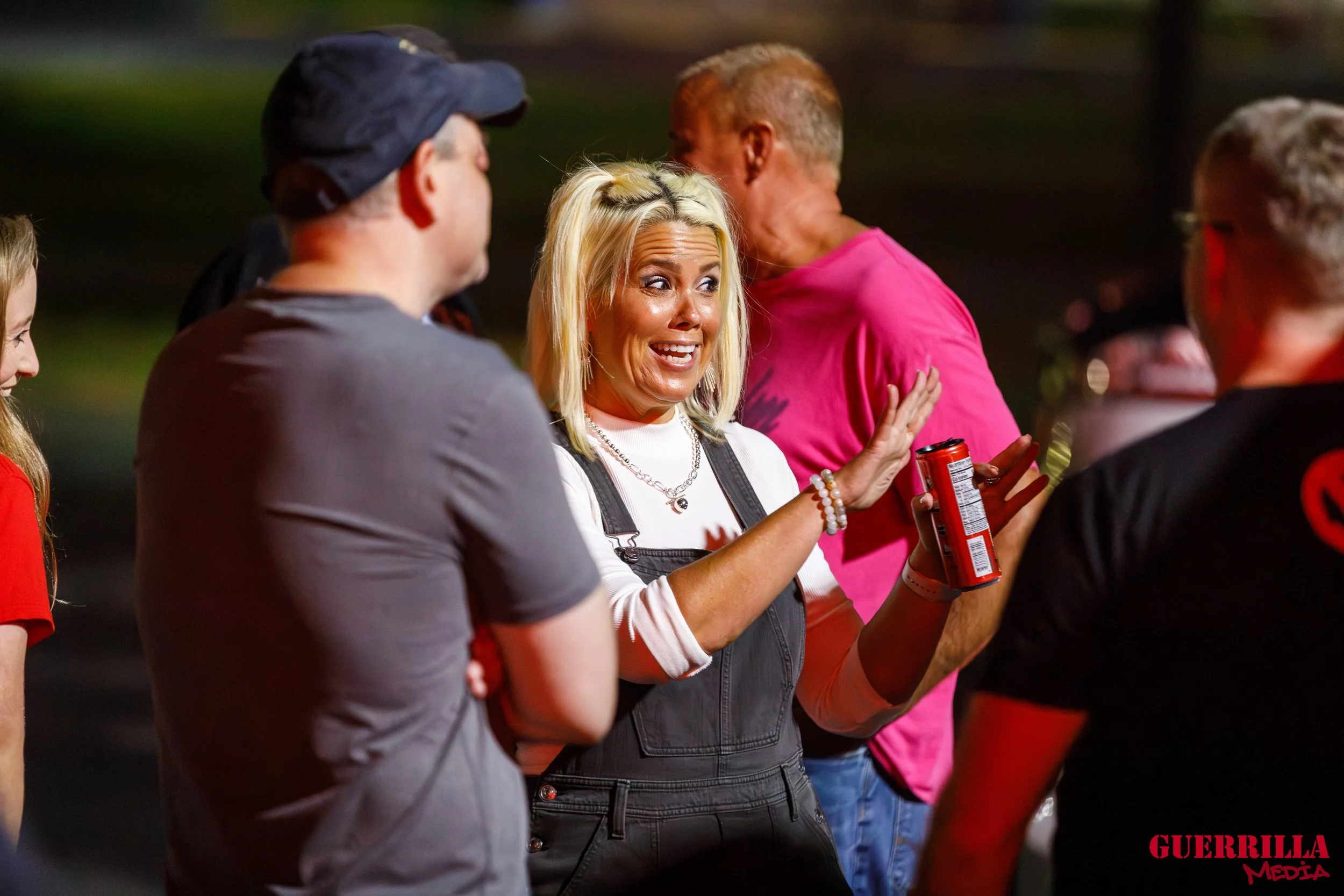 A woman with blonde hair, wearing a white shirt and dark overalls, appears to be upset or distressed as she talks to a group of people at an outdoor gathering at night. She is holding a red can of drink. The group is engaged in conversation, with som