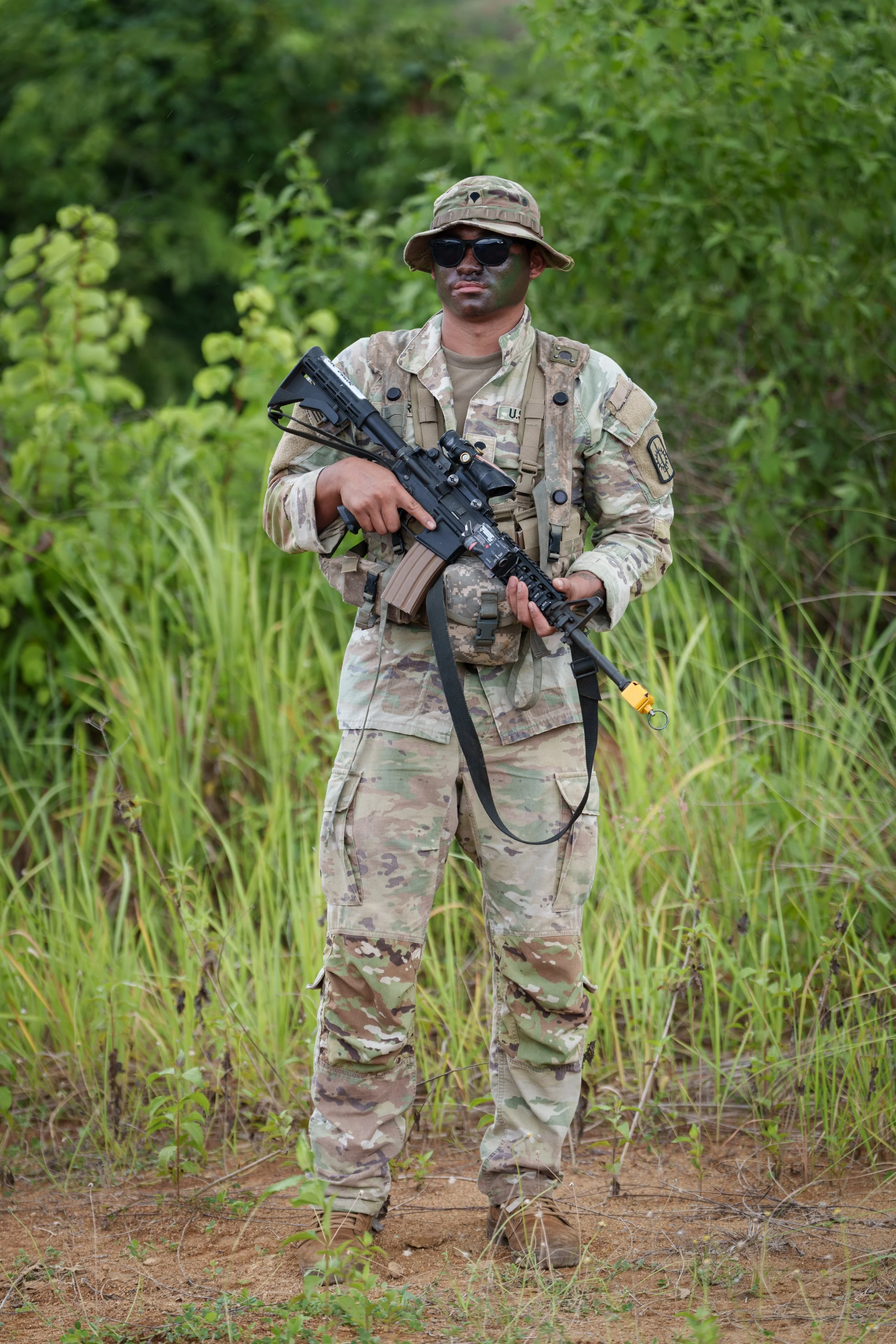 A soldier in camouflage uniform holding a rifle, standing outdoors in a grassy area with green foliage.