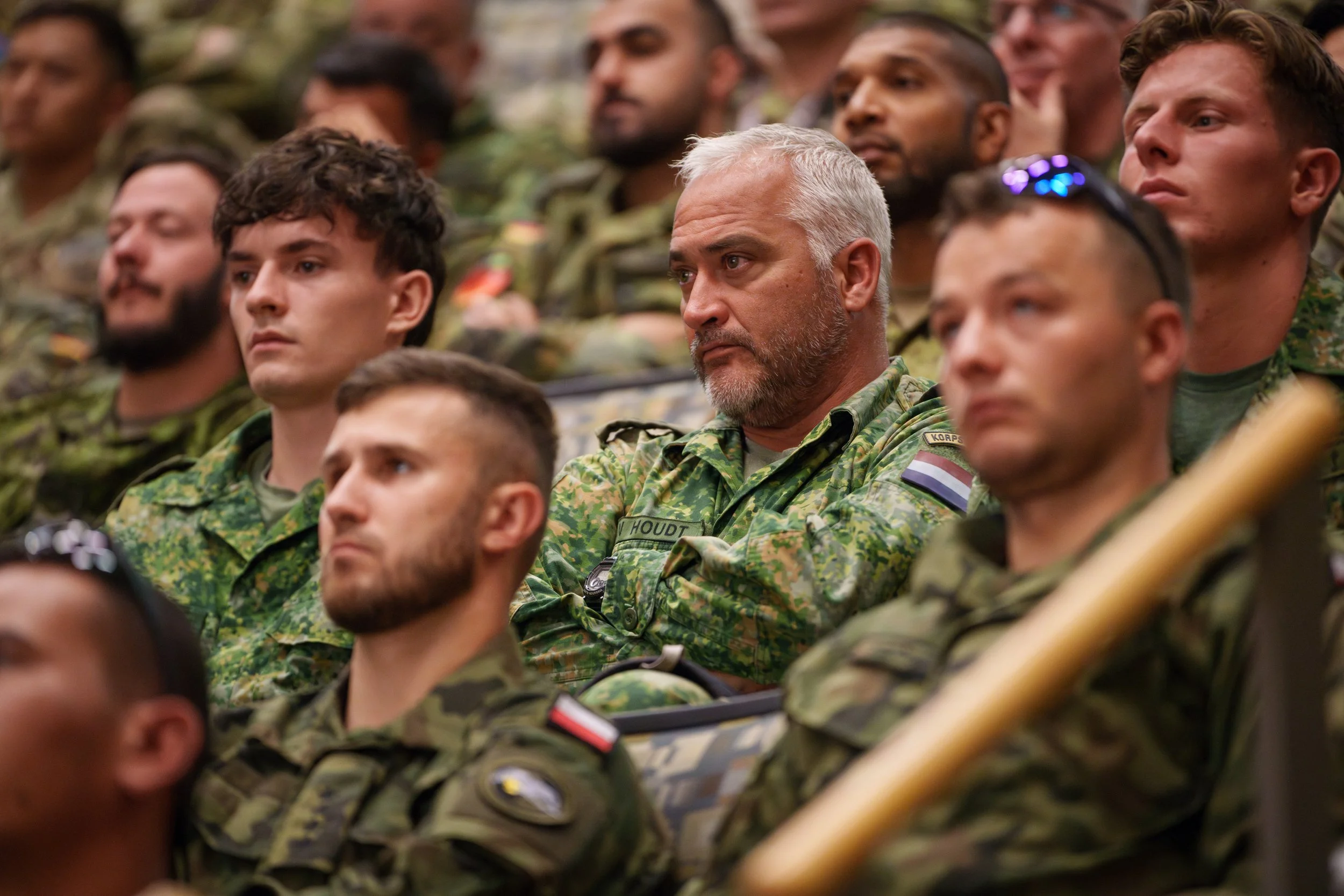 Group of soldiers in camouflage uniforms sitting and attentively listening at a military event or training session.