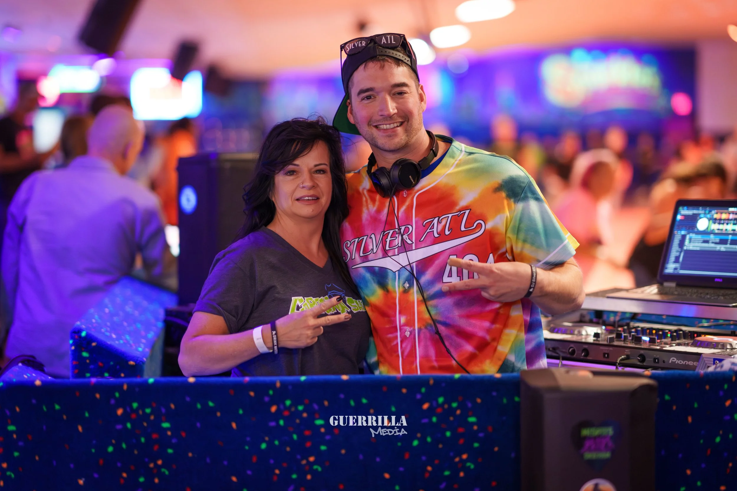 Two people, a woman with dark hair and a man wearing a colorful tie-dye shirt, are posing together at a party or nightclub. The woman is making a peace sign with her fingers. The man is smiling, pointing at the woman, and has headphones around his ne