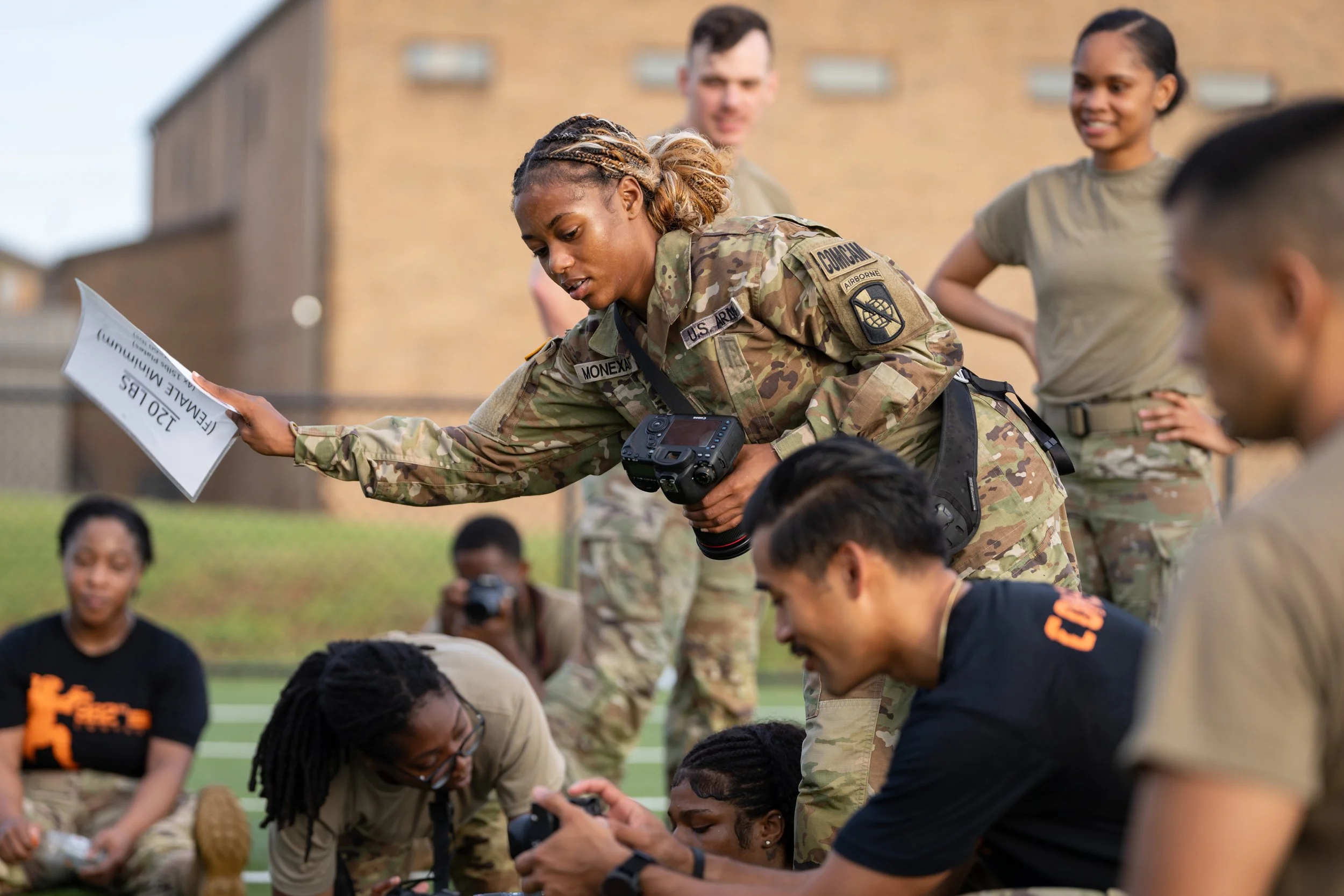 A diverse group of people, including military personnel and civilians, on a sports field during an outdoor activity or training involving navigation or map reading.