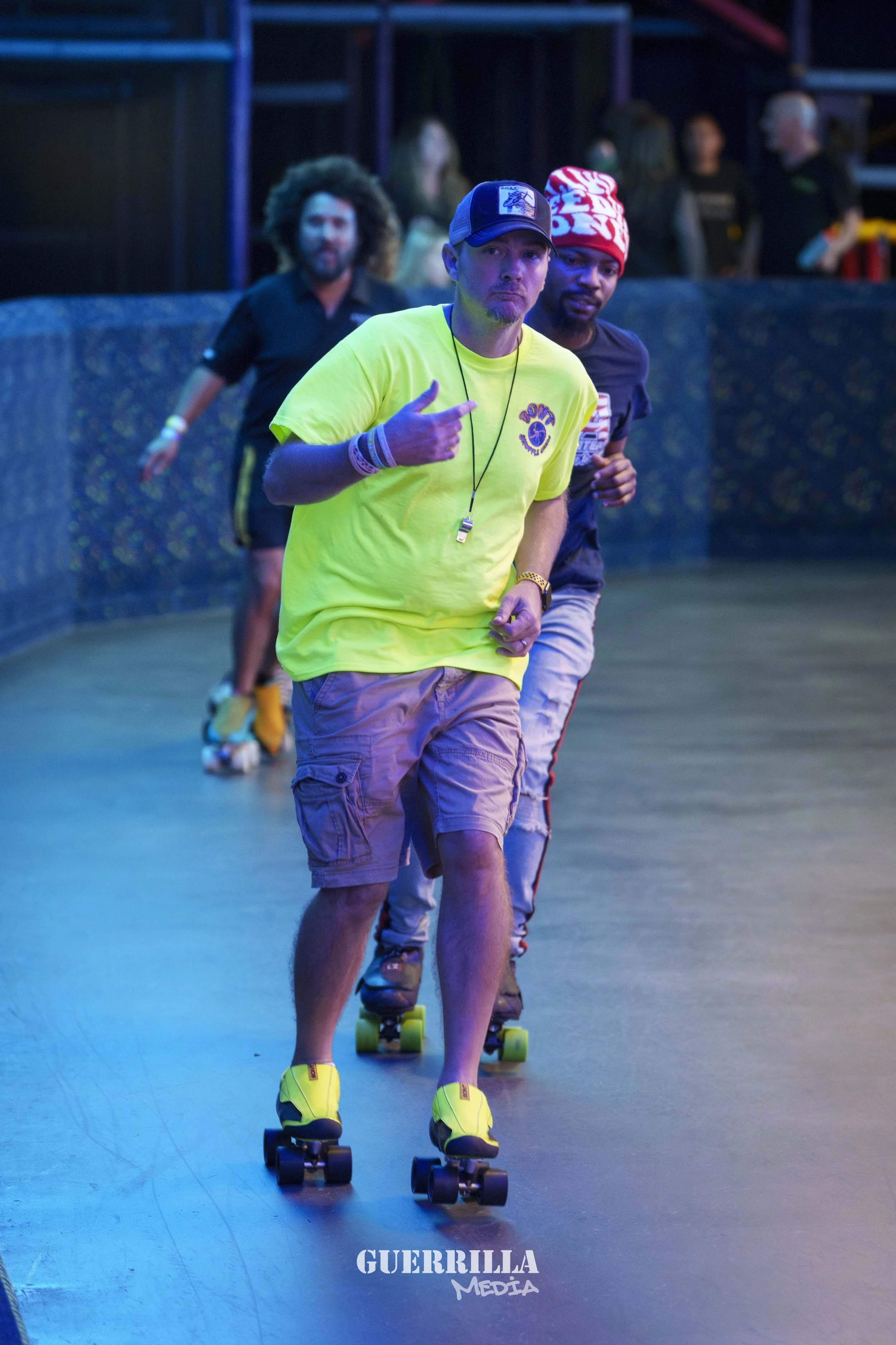 Three people roller skating on a rink. The person in front is wearing a bright yellow shirt, beige shorts, and yellow roller skates. The second person is wearing a dark shirt, gray pants, and roller skates. The third person is in the background, with