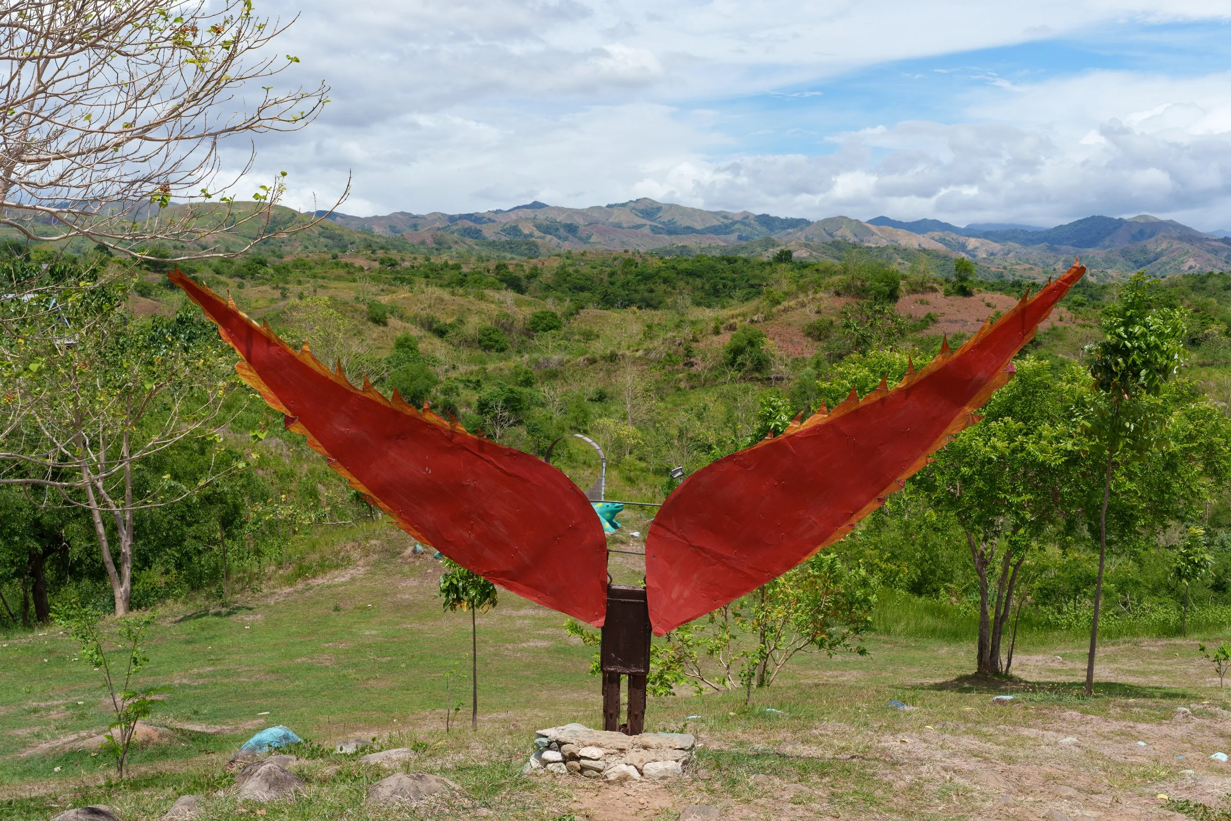A sculpture of large red butterfly wings with jagged edges, mounted on a stand, in a green outdoor landscape with trees and mountains in the background.