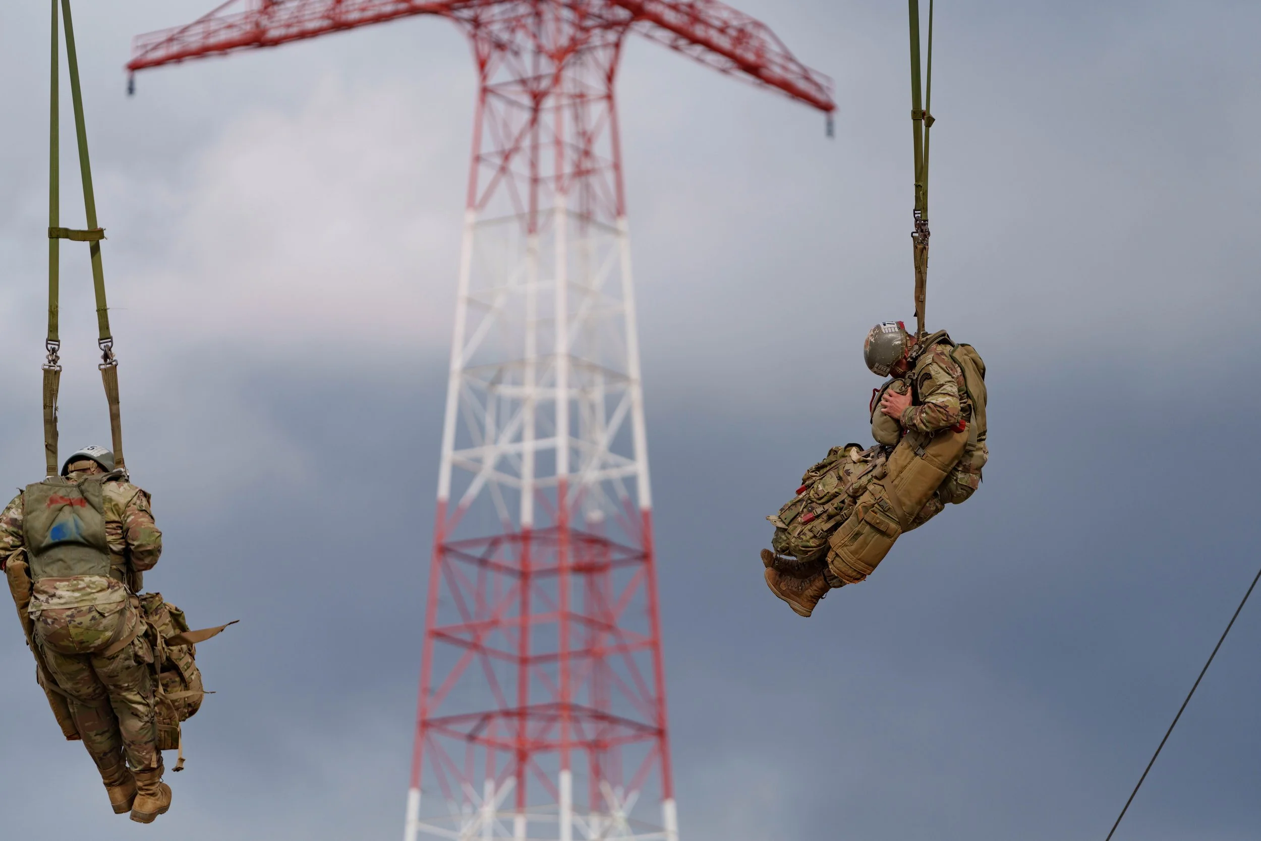Two soldiers in military gear suspended on a zip line, with a red and white communication tower in the background under cloudy skies.