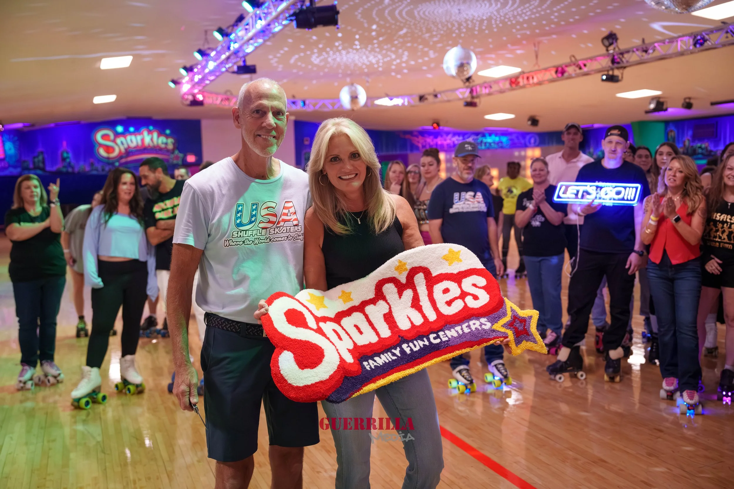 A woman and a man holding a Sparklers Family Fun Center sign at an indoor roller skating rink, with people roller skating in the background and colorful lighting.