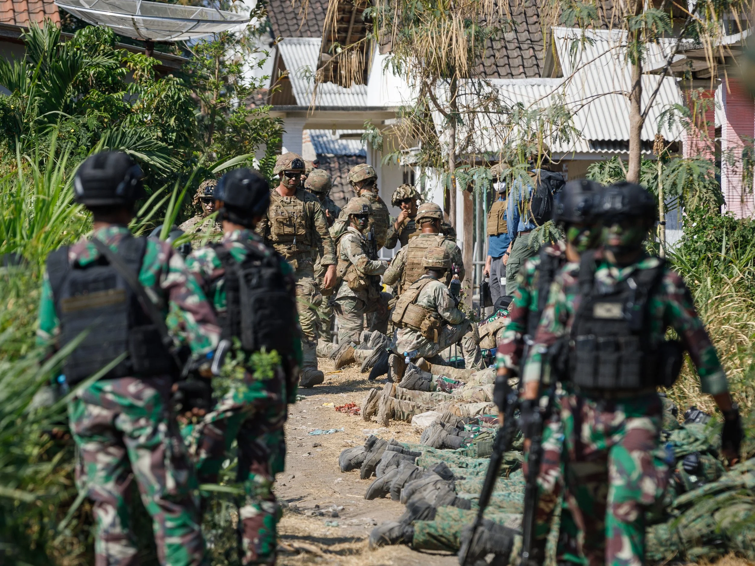 Military personnel gathered and kneeling on a dirt path in front of houses, with some soldiers standing and others kneeling with their weapons, surrounded by greenery.