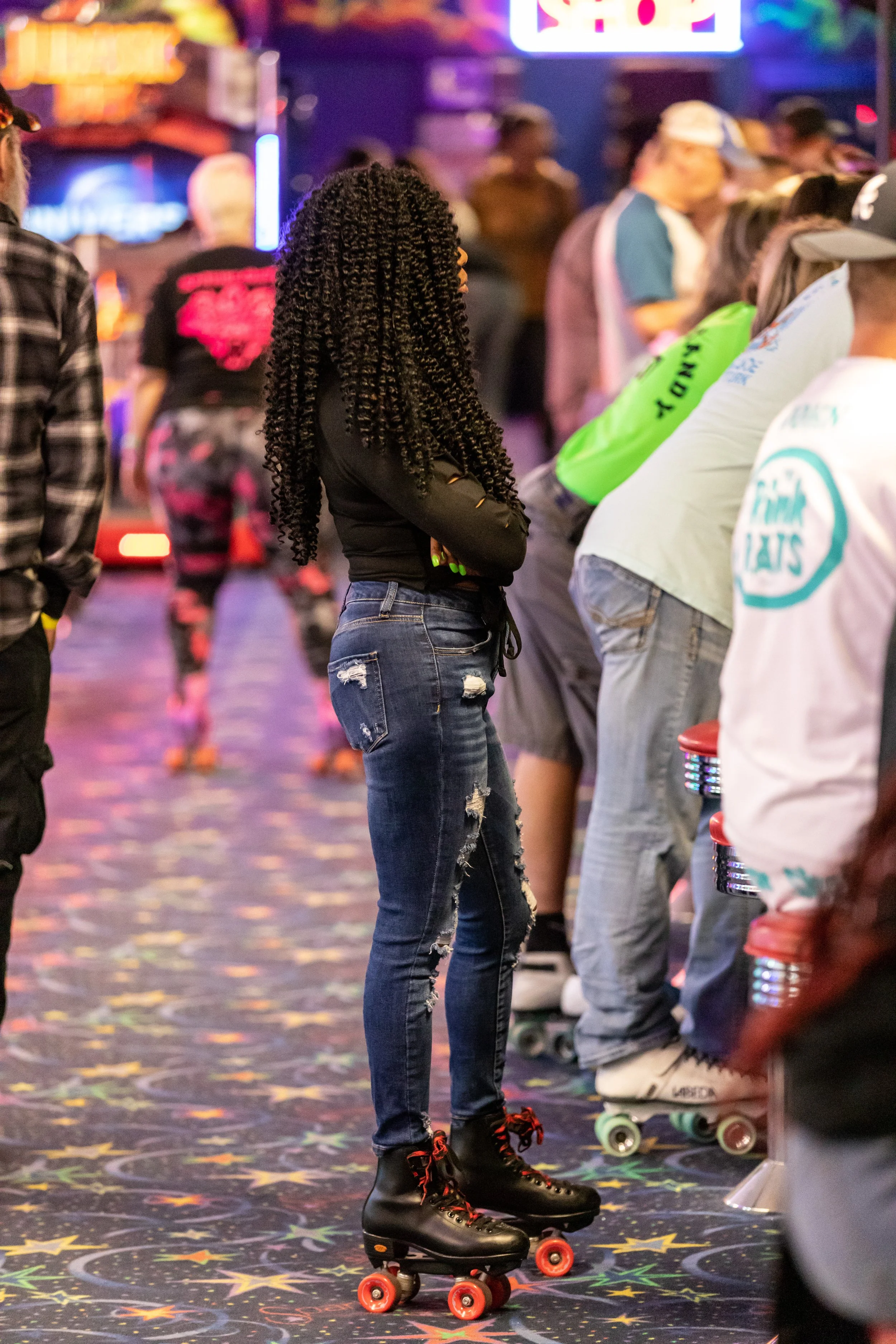 A woman with long, curly hair, wearing a black top and ripped jeans, is roller skating inside an arcade or entertainment center with neon lights and other skaters around her.