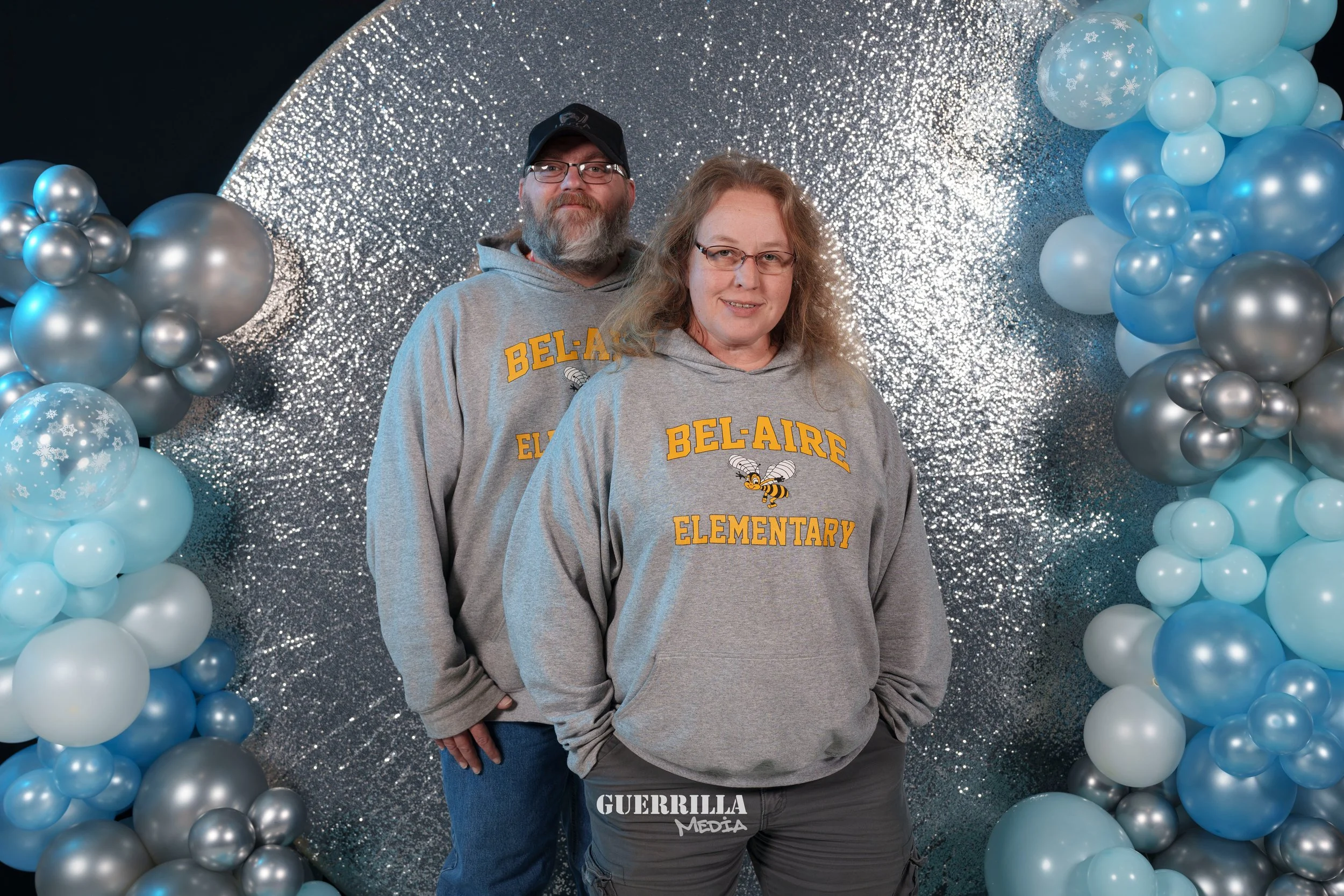 Two adults wearing gray sweatshirts with 'Belaire Elementary' and a bee logo, standing in front of a silver glittery backdrop surrounded by blue and silver balloons.