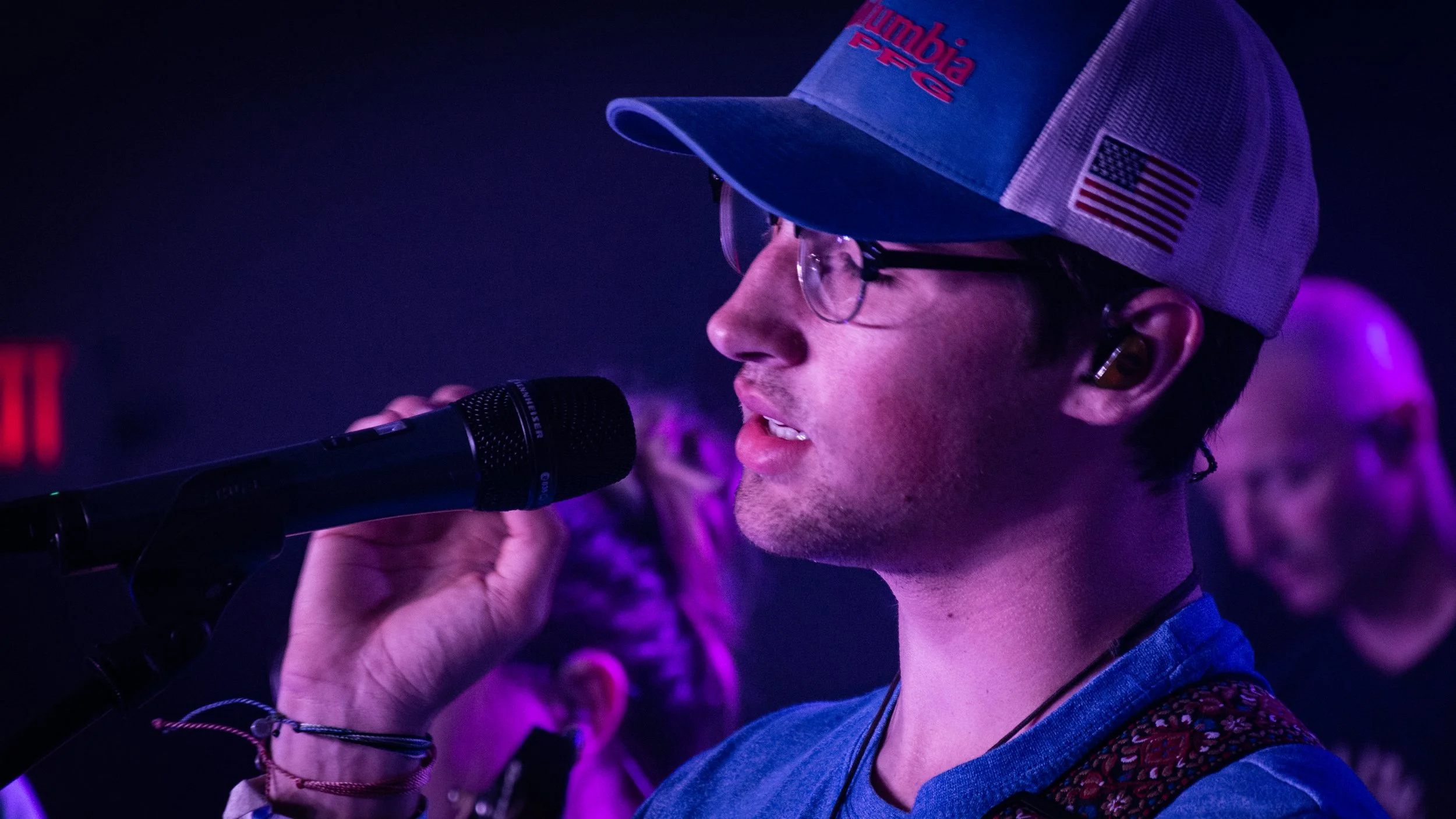 A young man with glasses and earbuds, wearing a trucker hat with an American flag and 'umbia PROS' text, speaking into a microphone in a dimly lit, purple-lit environment.