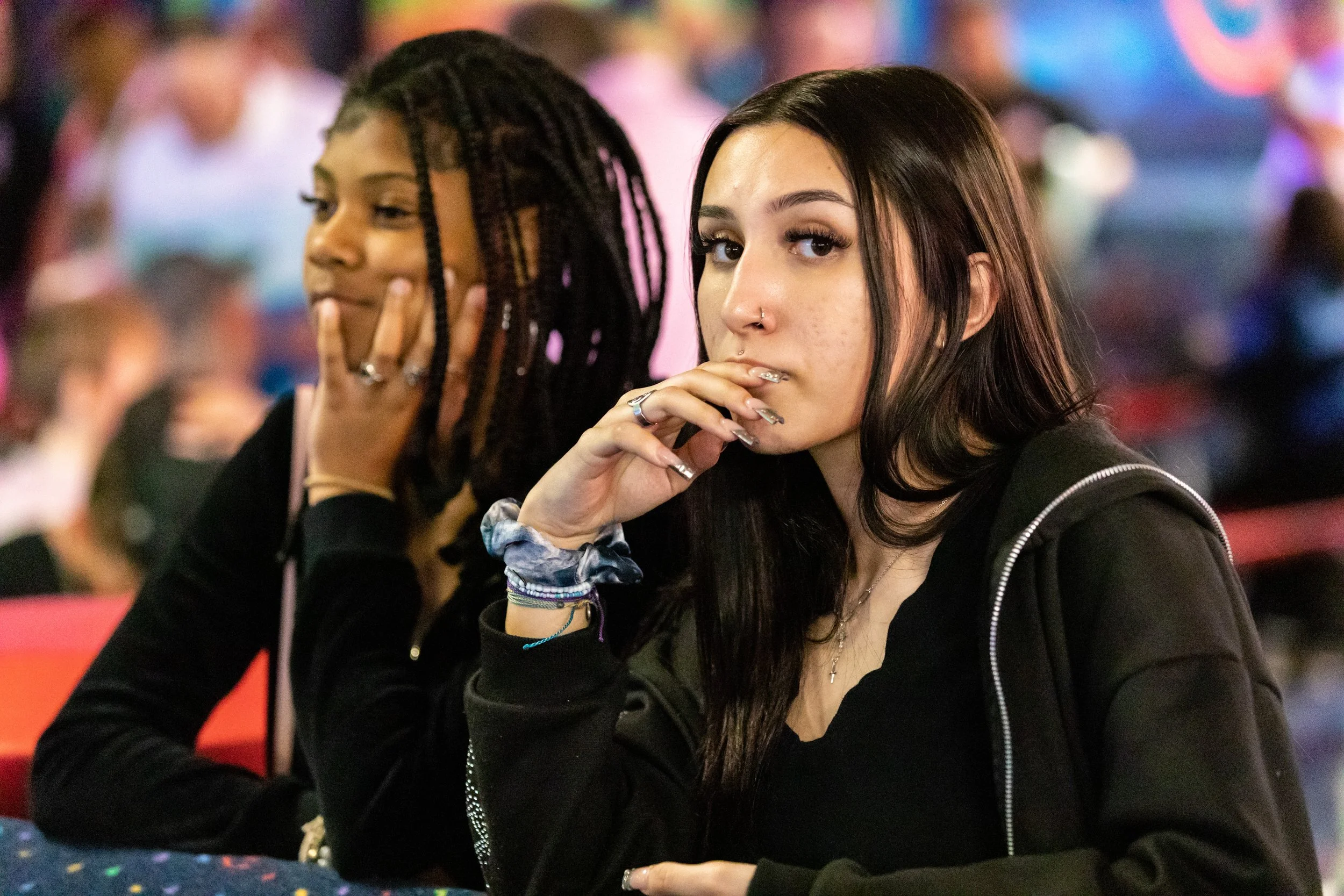 Two young women sitting at a table in a crowded setting, one with her hand on her face and the other resting her hand near her mouth, both with serious expressions.