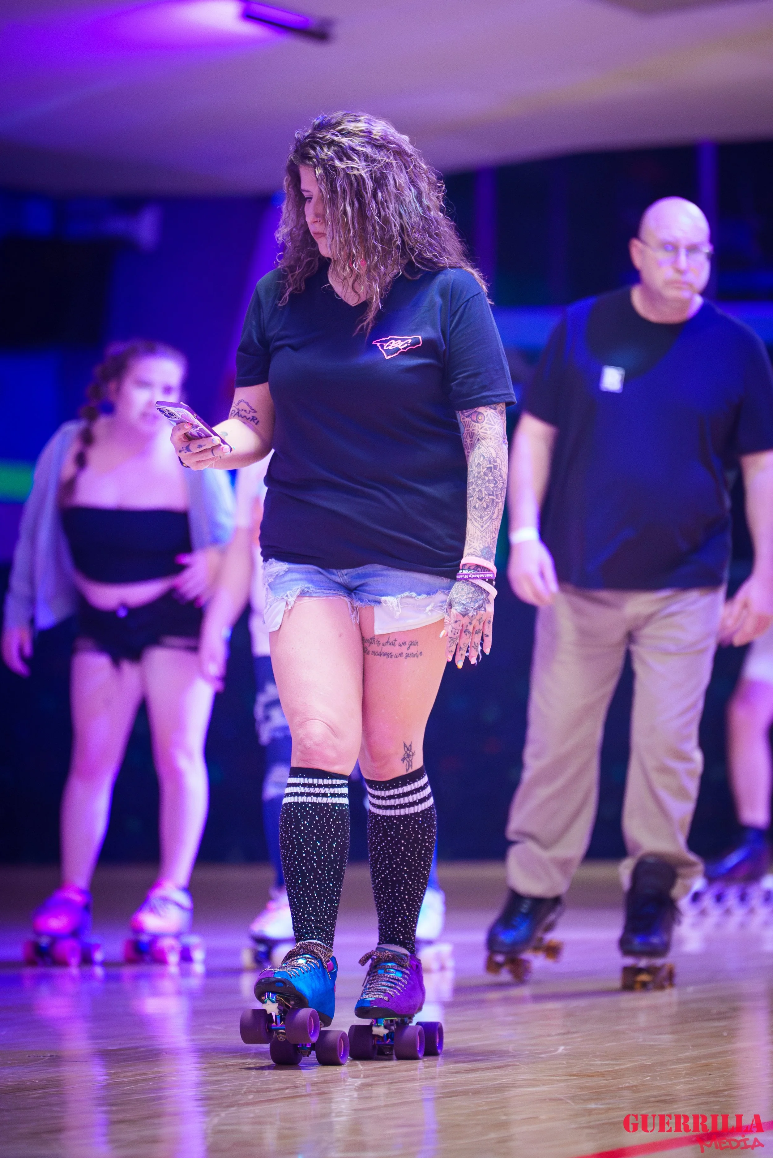 A woman with curly hair wearing a black t-shirt, denim shorts, black knee-high socks with white stripes and dots, and roller skates, is looking at her phone. She has tattoos on her arms and one on her leg. Behind her, a man with glasses in a black t-