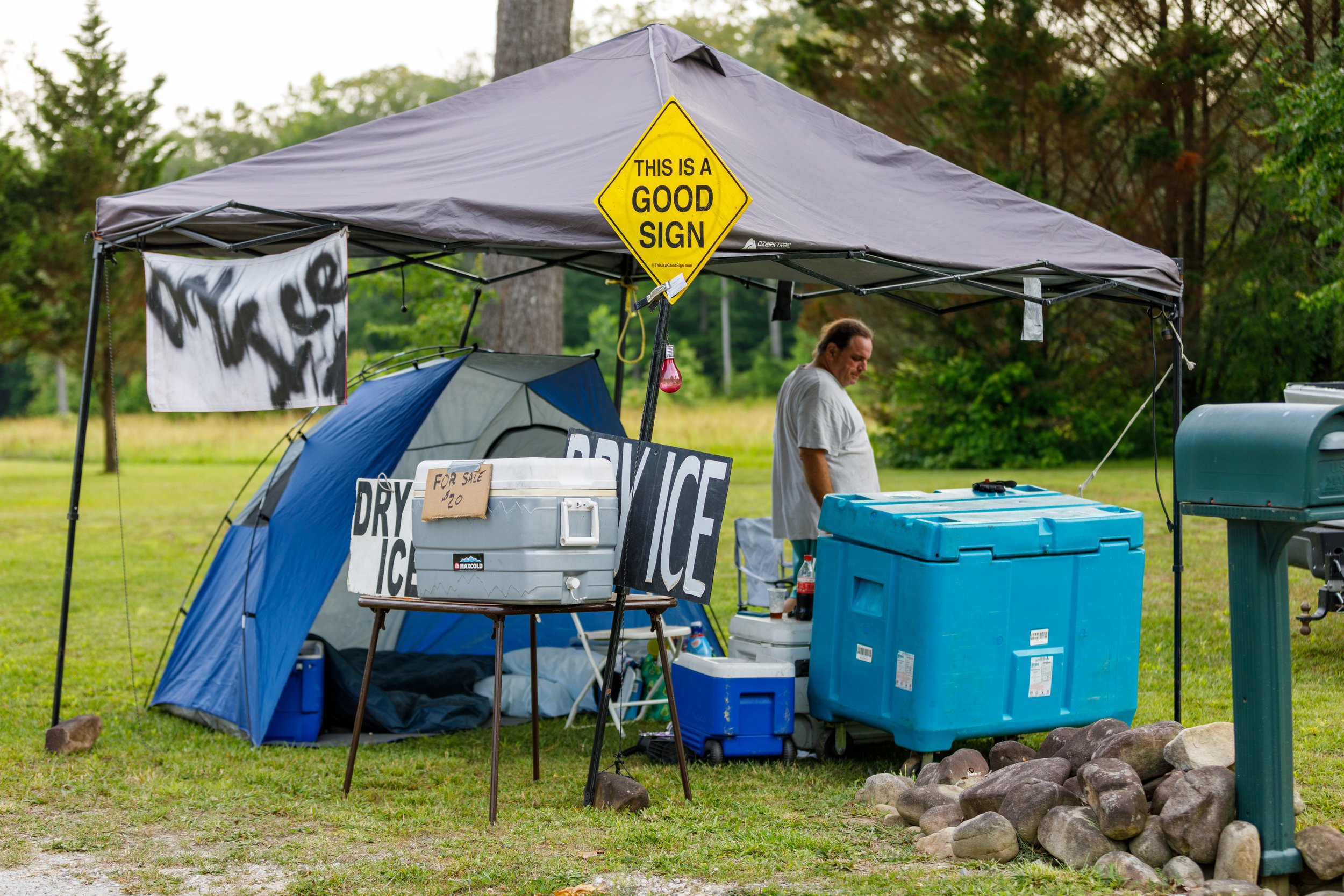 A man standing under a gray canopy tent next to a large blue cooler and a mailbox, surrounded by signs advertising dry ice and a good sign, with a small blue and gray tent, gravel, rocks, and trees in the background.