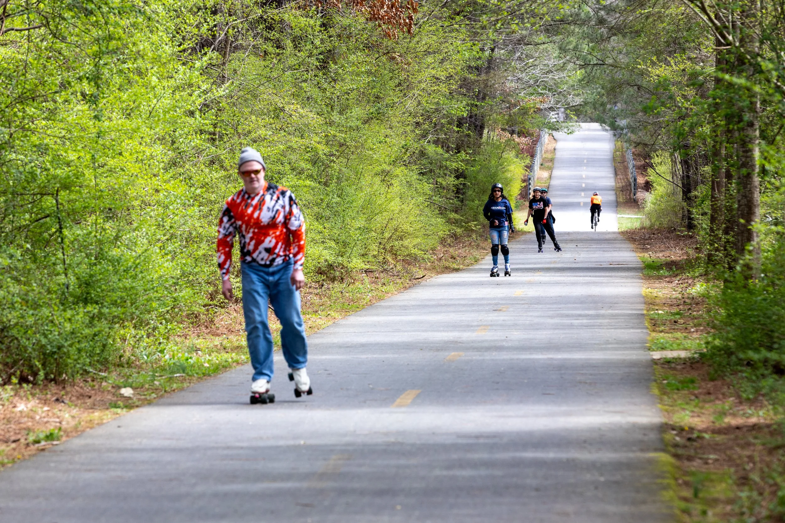 People roller skating and skateboarding on a paved path in a green wooded park.