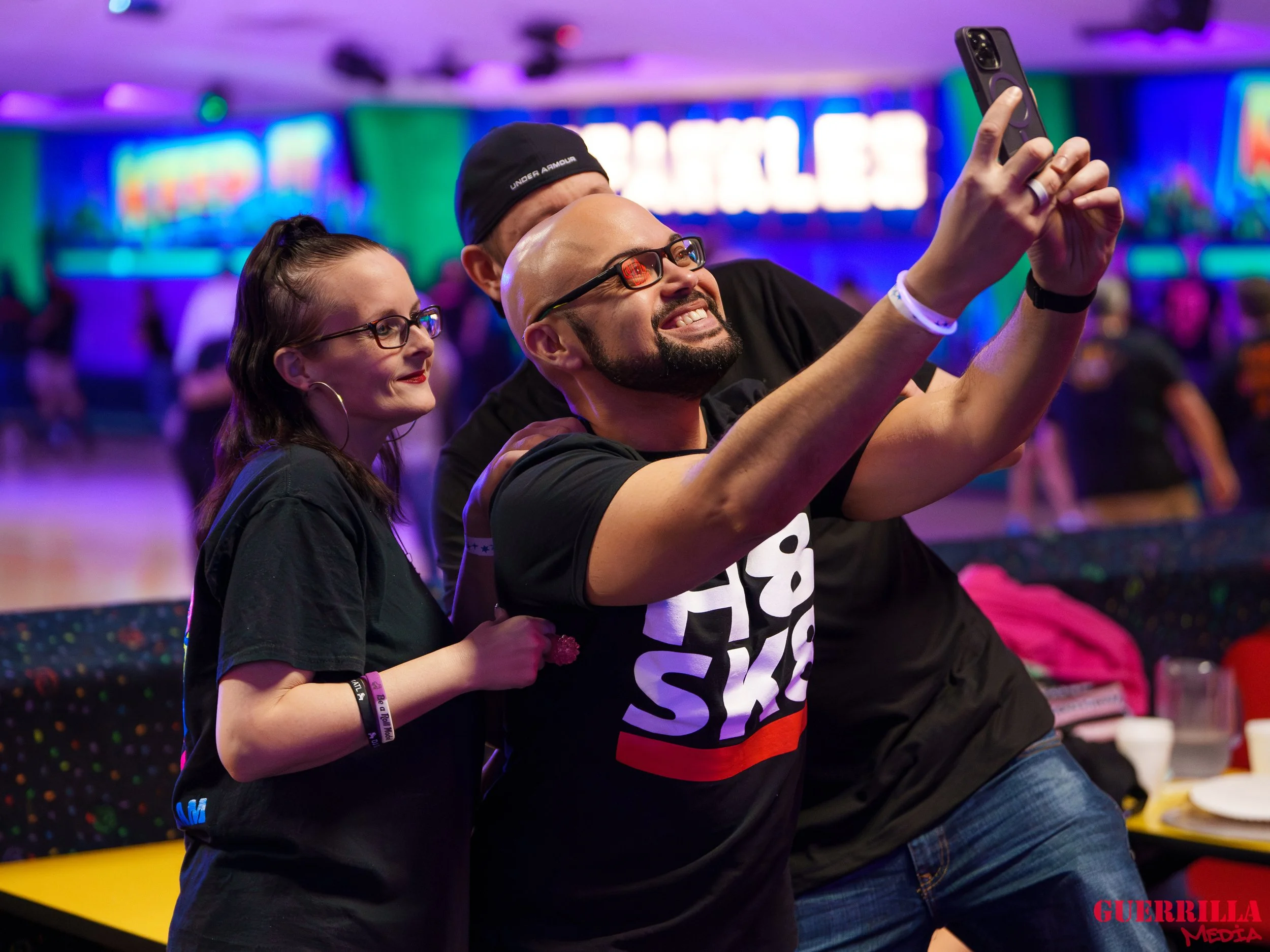 Three people taking a selfie at a neon-lit event or party. The man in the middle is smiling widely, holding a smartphone, with two others beside him. The background shows a vibrant, colorful setting with blurred lights and a lively atmosphere.