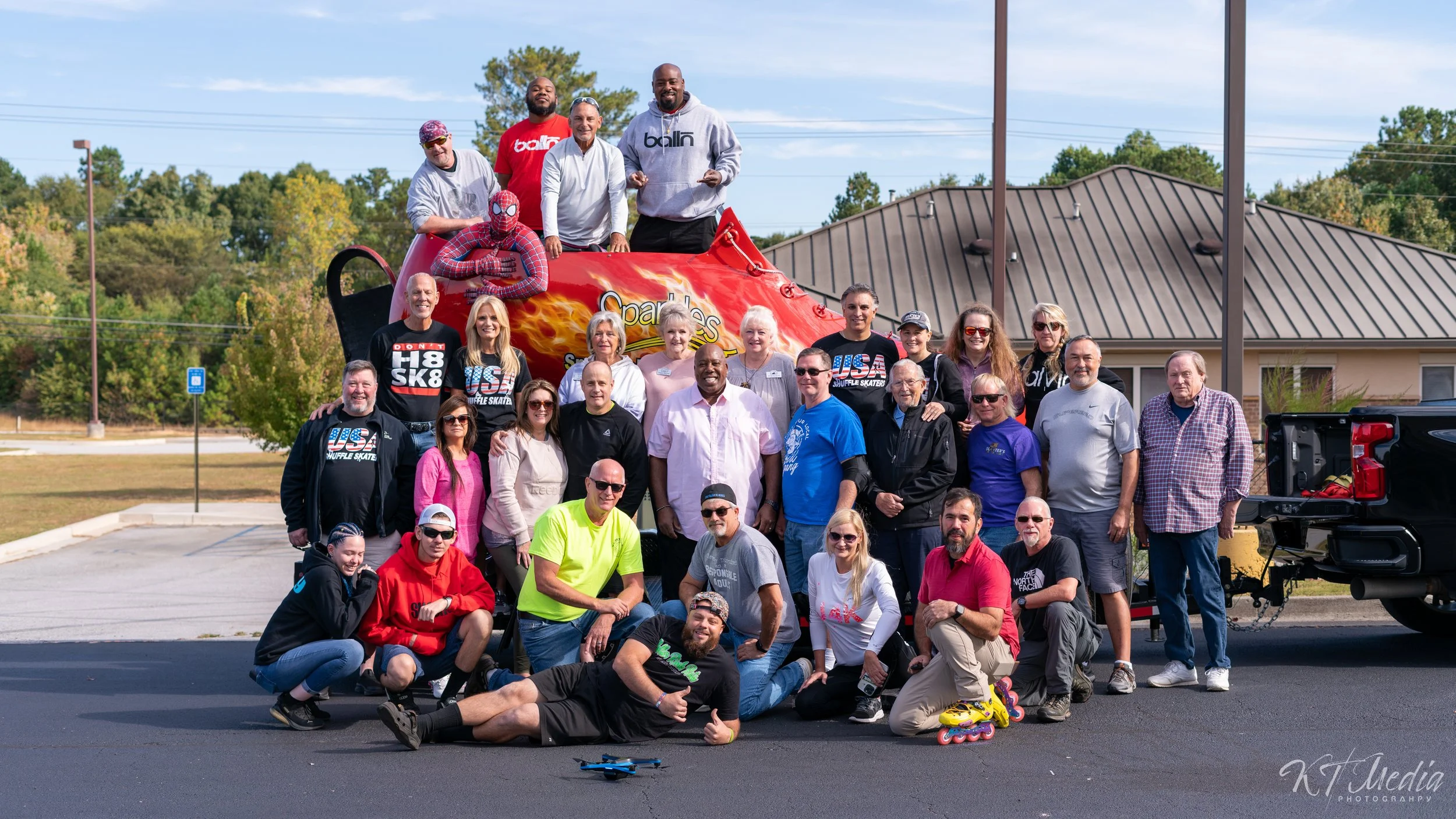 A large group of people gathered around a giant pair of roller skates with a hot sauce bottle design, with some sitting, kneeling, or standing in front of the skates. The background includes a building, trees, and a parking lot.