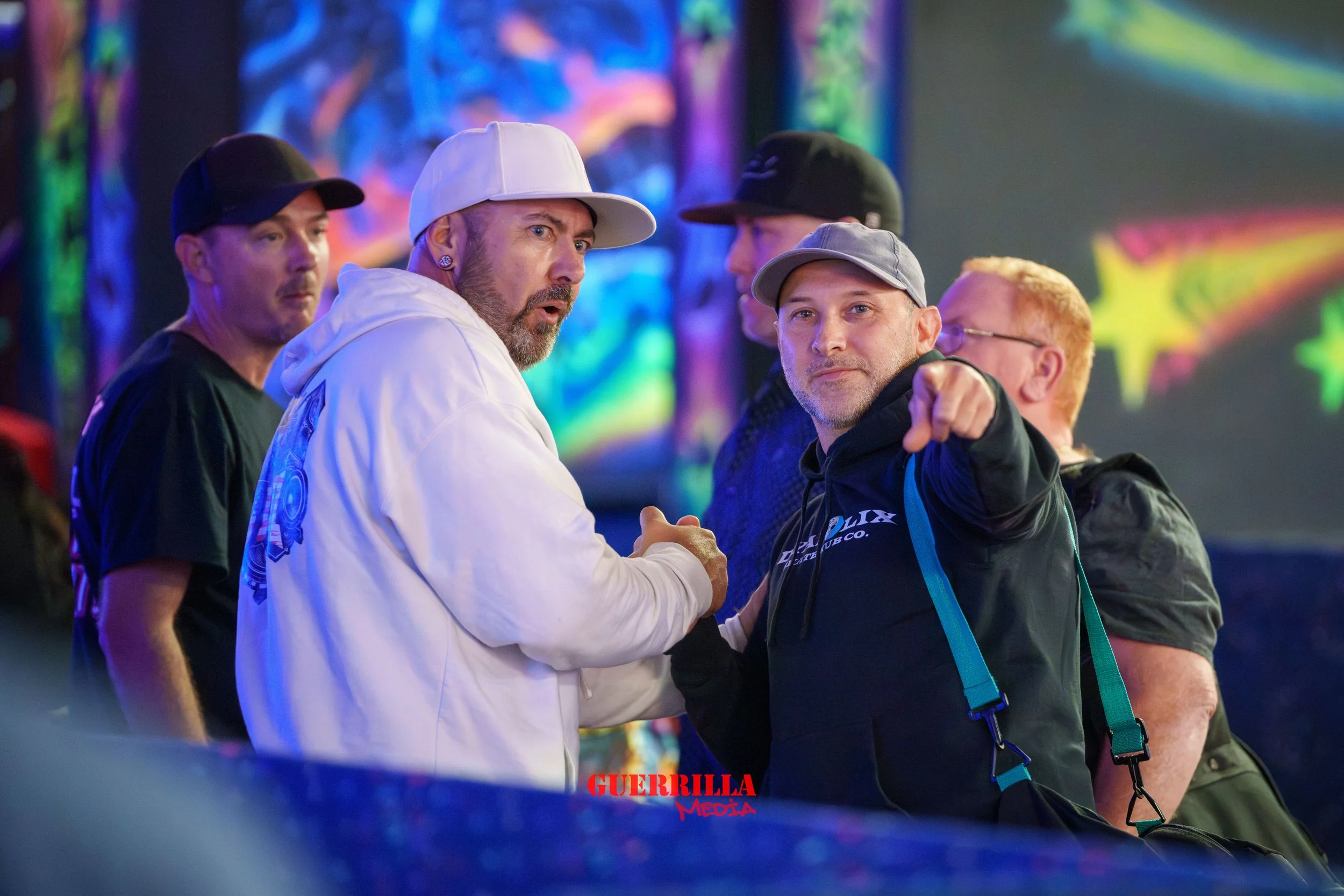 A group of five men in a colorful, illuminated setting. One man in a white hoodie and white cap points directly at the camera while another man in a black hoodie and gray cap shakes his hand. The others look on, with some wearing caps and glasses.