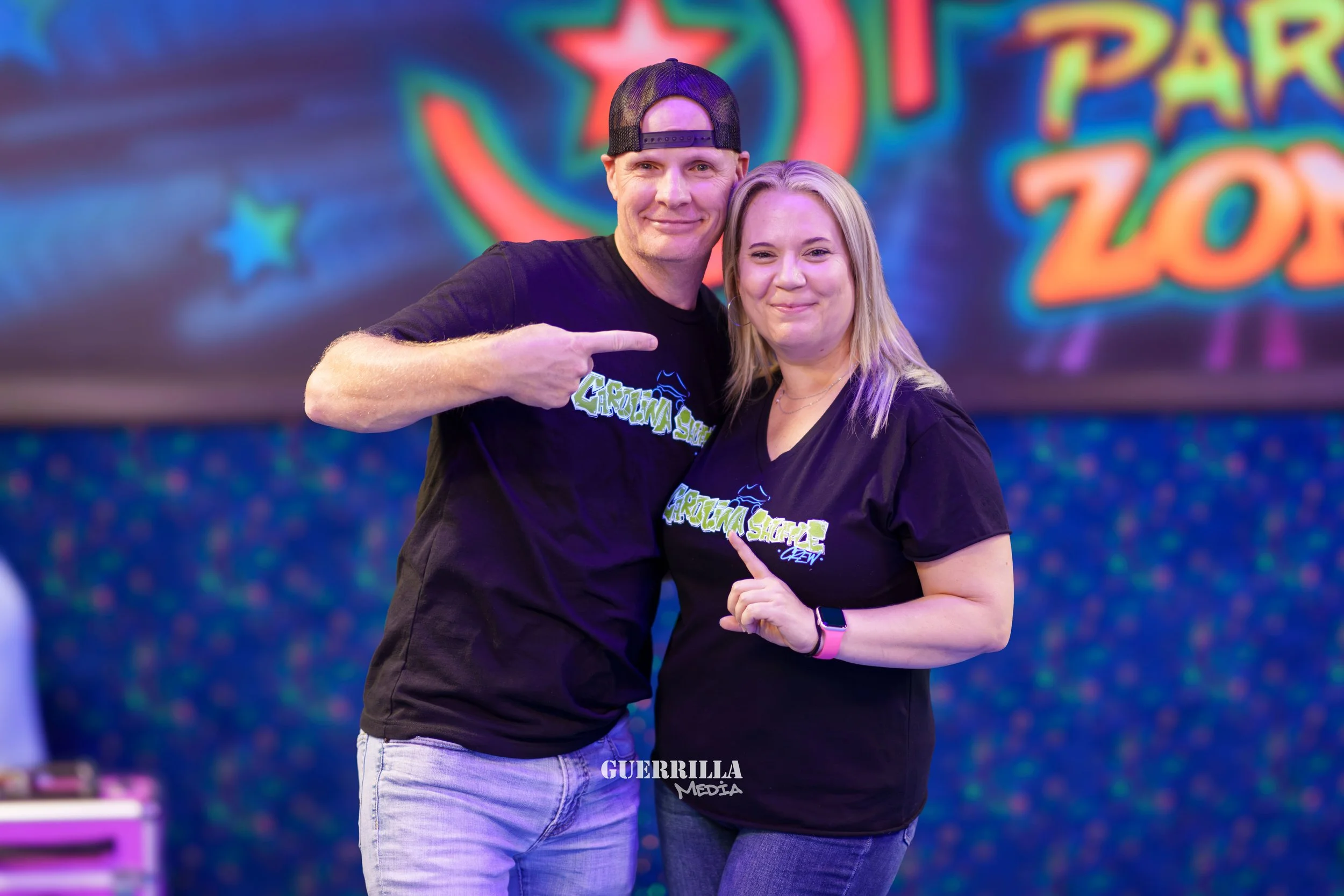 A man and woman standing together at an event, both wearing black T-shirts with green and blue text that says ‘GROWIN SHARES CREW’. The man is pointing at the woman, and they both are smiling. The background features colorful, blurred graphics and th
