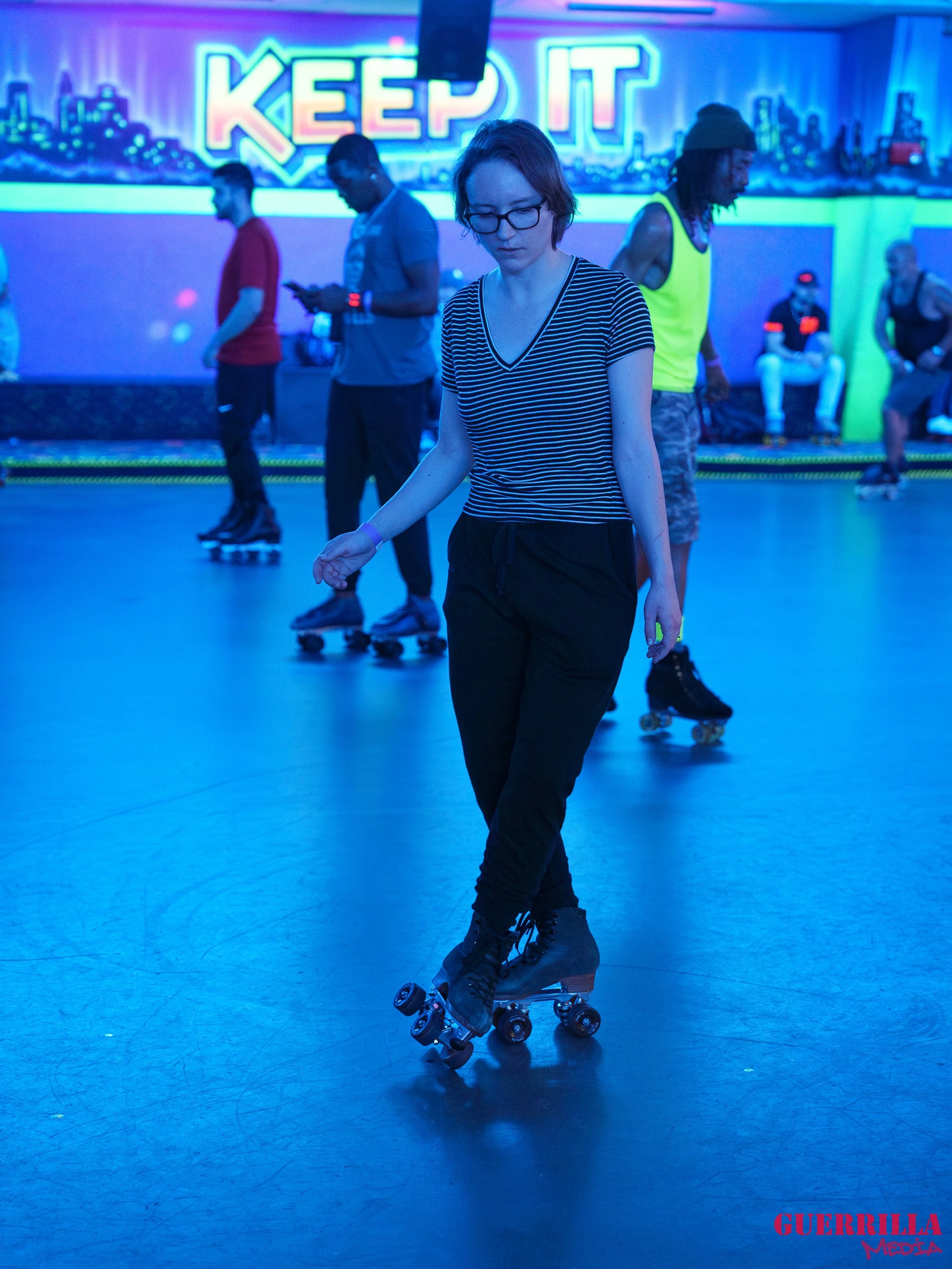 A young woman is roller skating in an indoor rink with blue lighting. She is wearing a black and white striped shirt, black pants, and black roller skates. In the background, people are also skating, and a colorful mural with the text 'KEEP IT' is vi